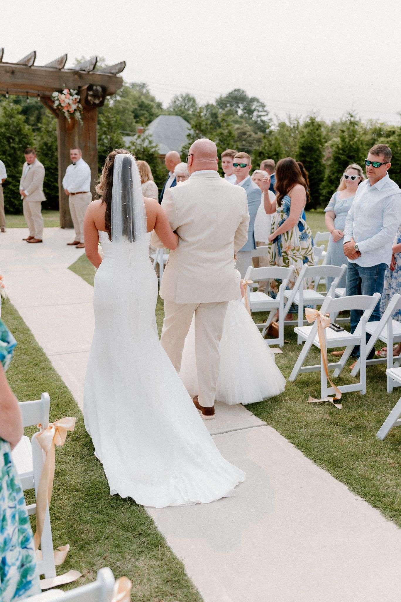 A row of wedding guests stand and face the center aisle as several people in white dresses walk down the outdoor pathway.