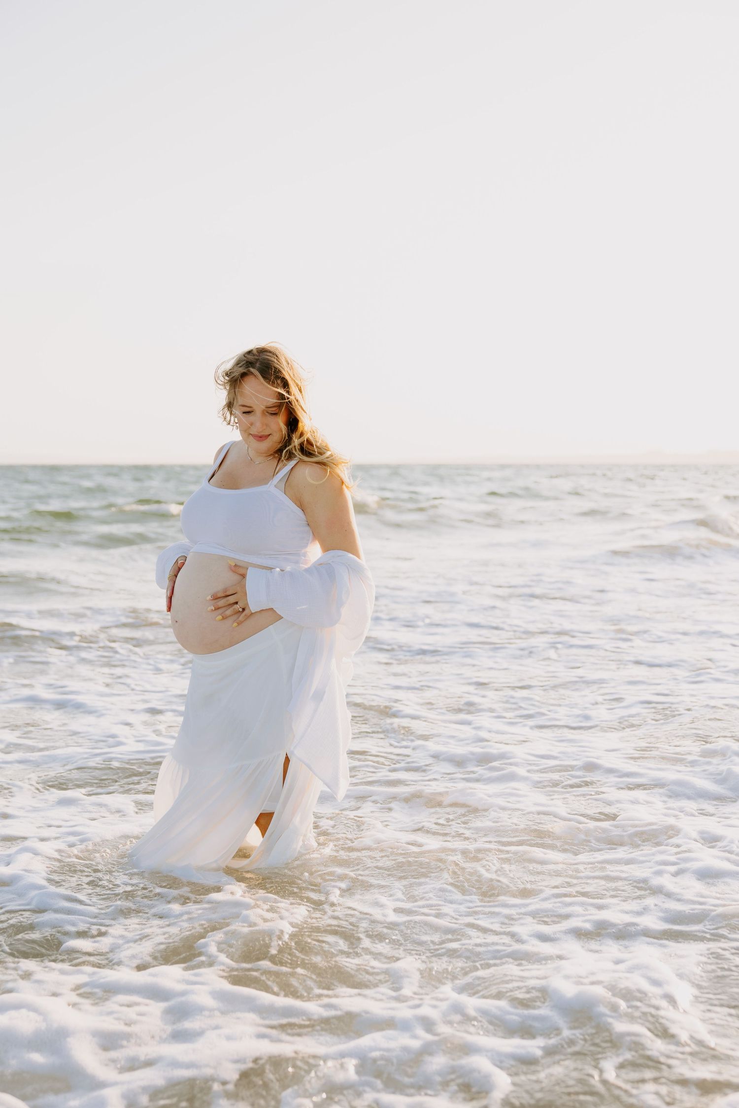 An expectant mother in a flowing white dress stands in gentle ocean waves.