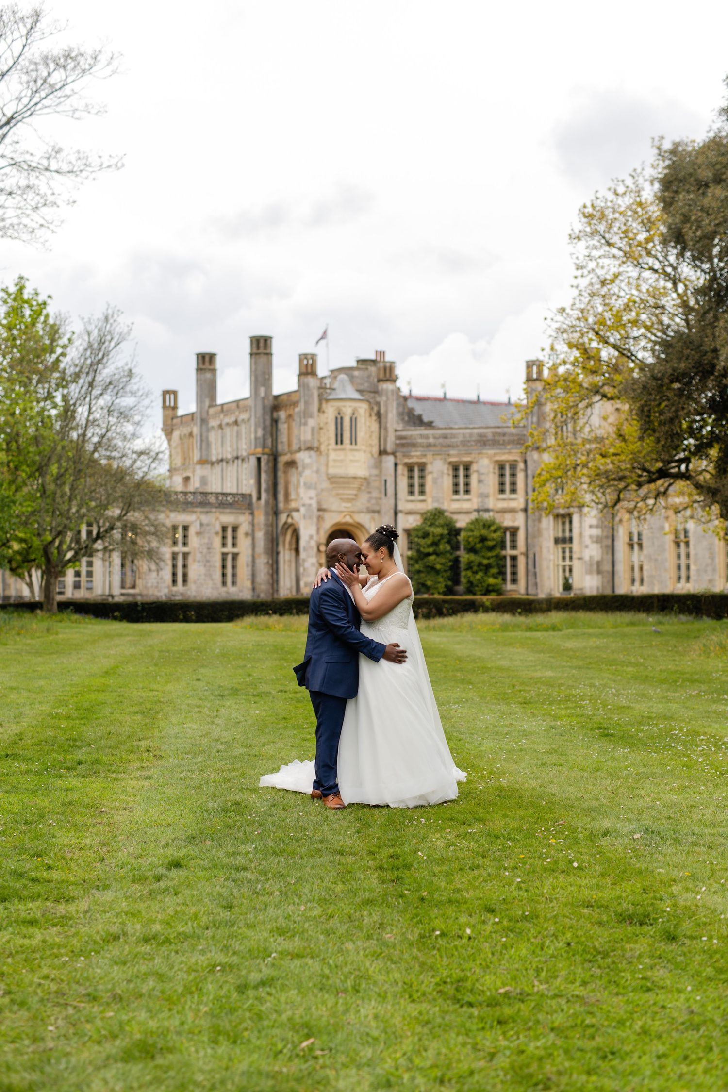 A romantic wedding couple embraces on a grand lawn in front of a stunning historic castle venue.