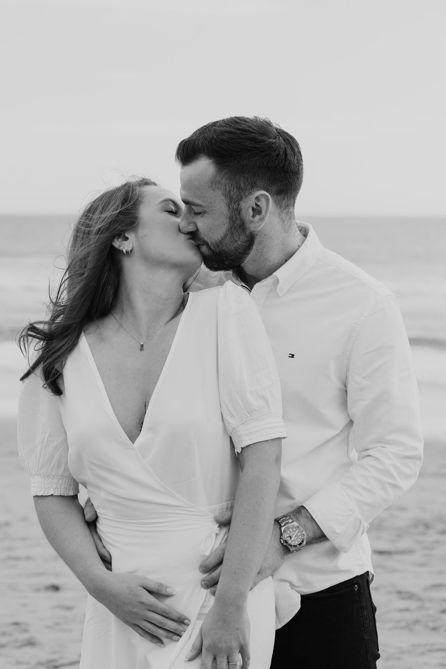 A romantic black and white photo of a couple kissing on the beach at sunset.