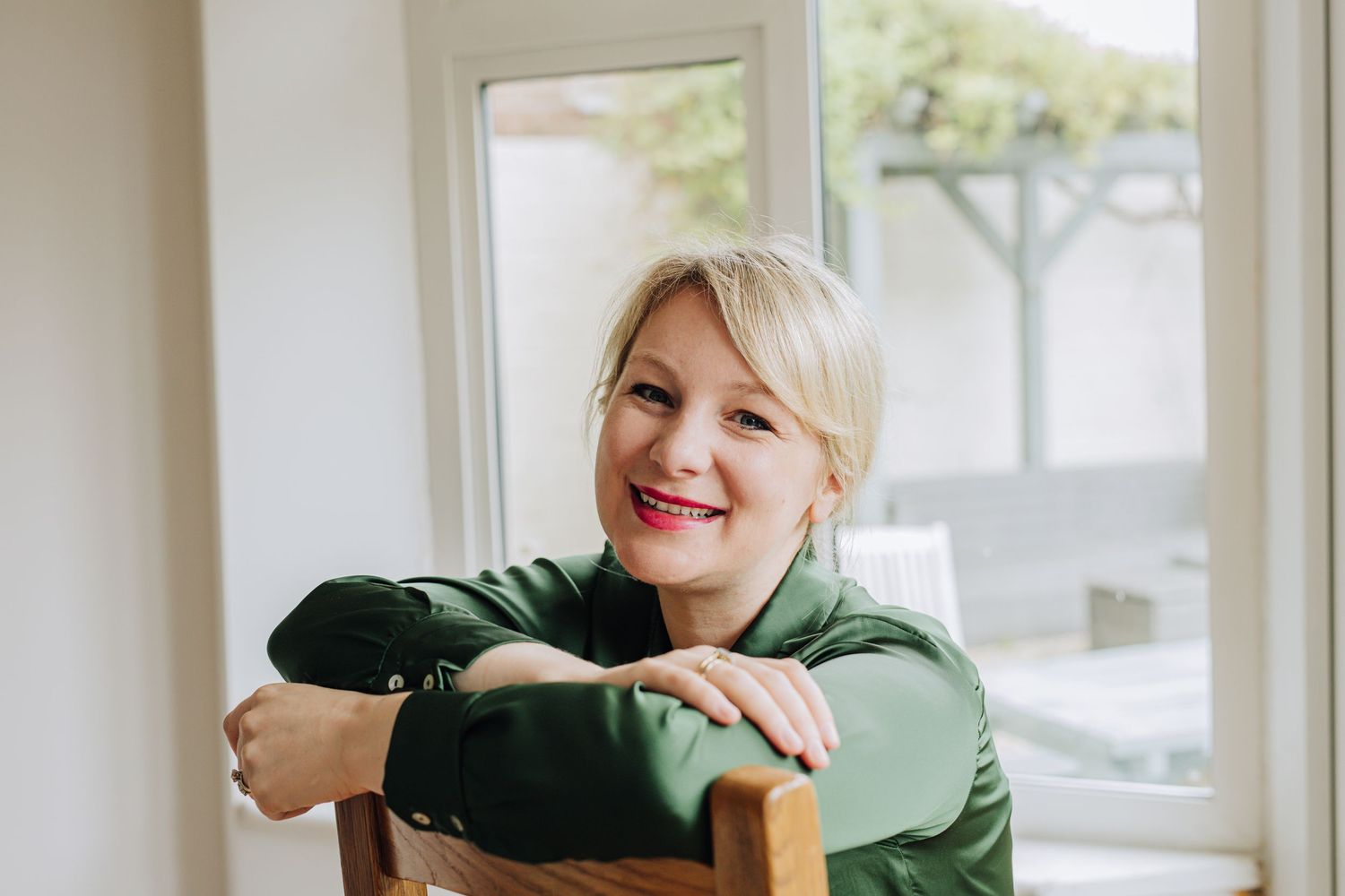Someone wearing a green sweater smiles while leaning on a wooden chair in a bright indoor setting.