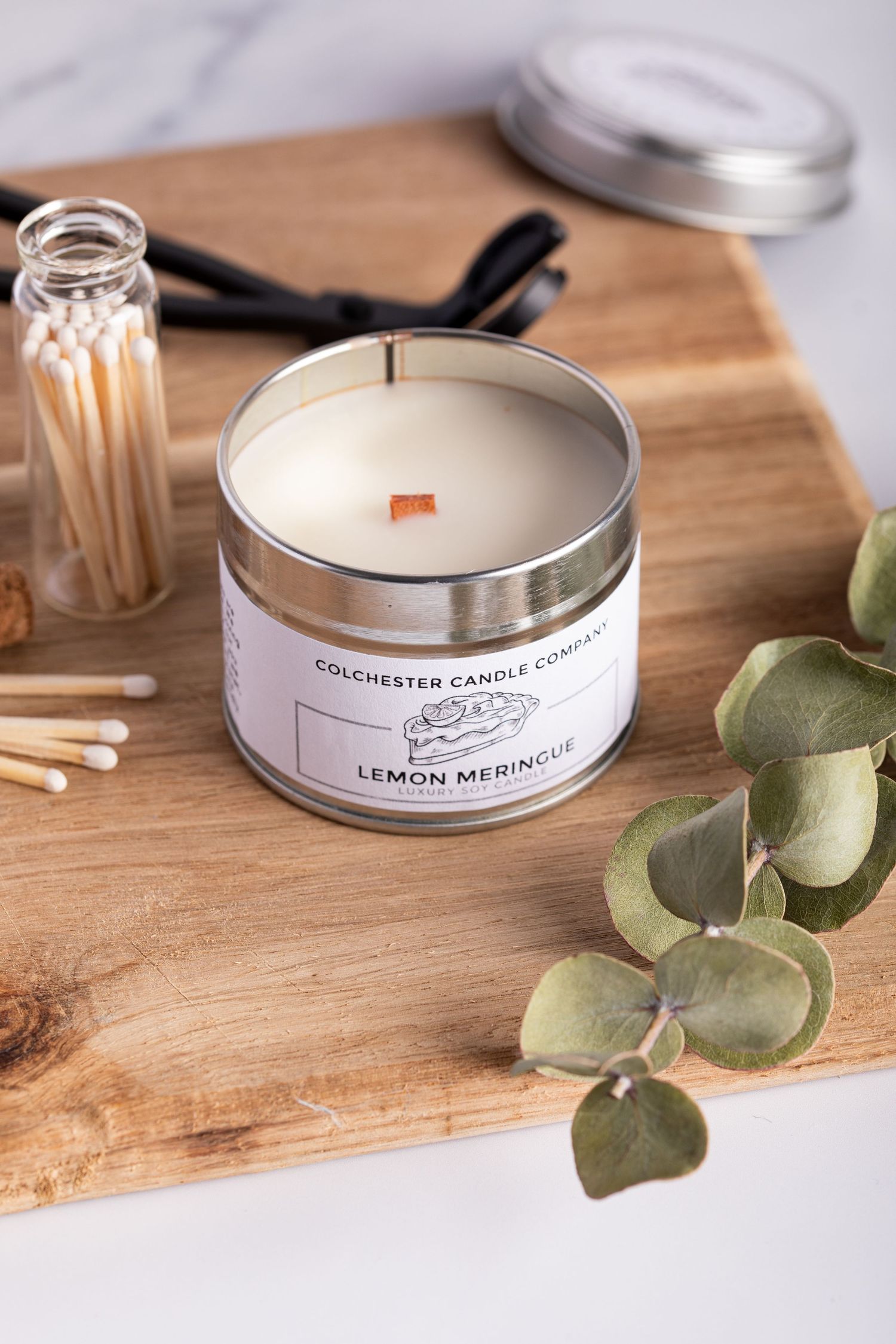 Wooden cutting board displays a lemon meringue scented candle in tin container with matches and eucalyptus leaves.