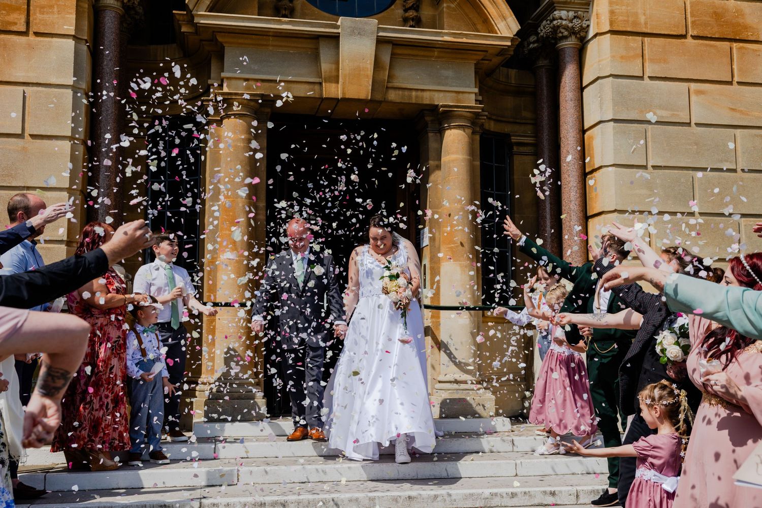Wedding guests shower newlyweds with confetti as they exit historic stone church steps.