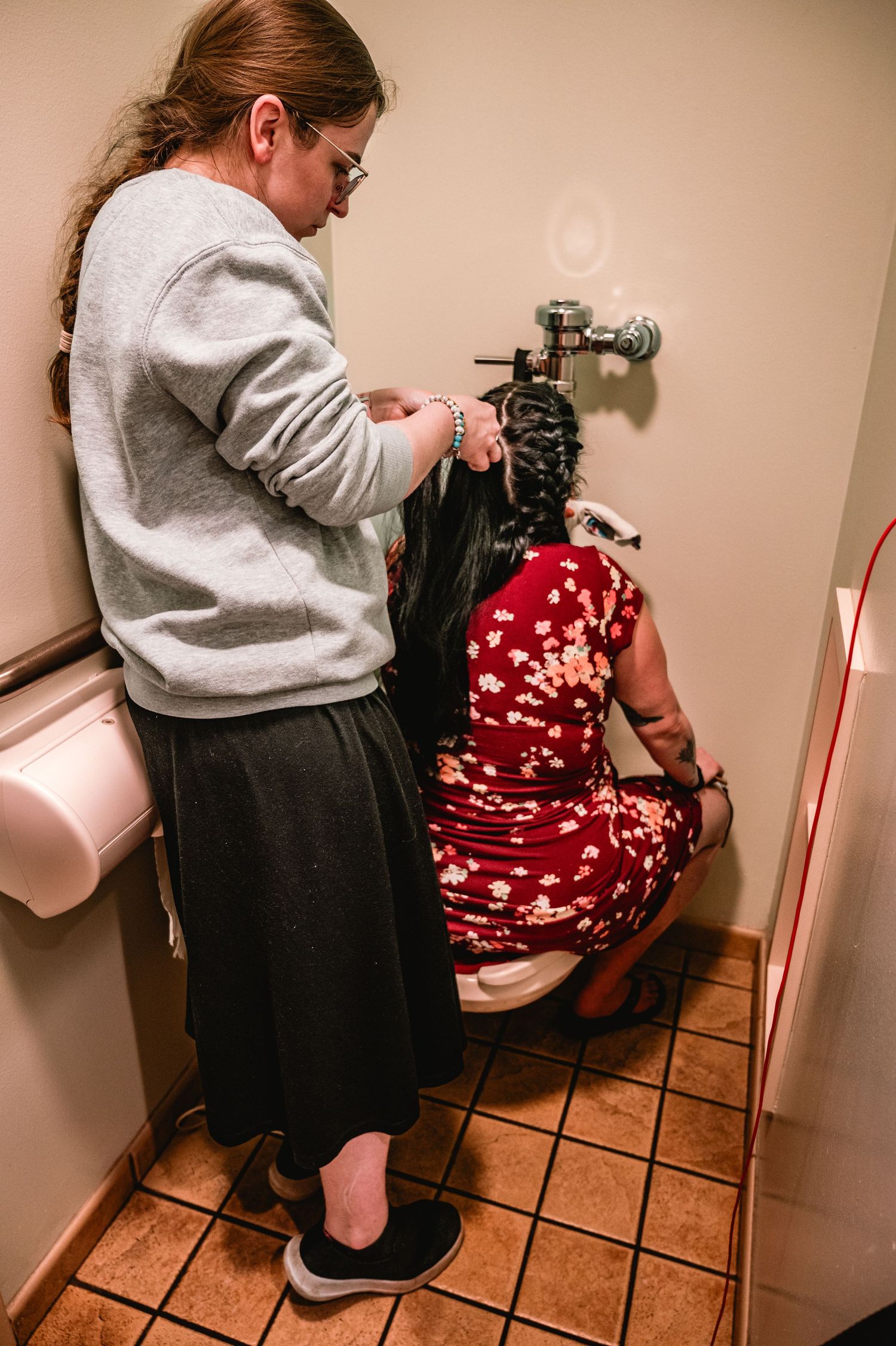 Person in gray sweatshirt helps style someone's hair in a small bathroom with terracotta tile flooring.