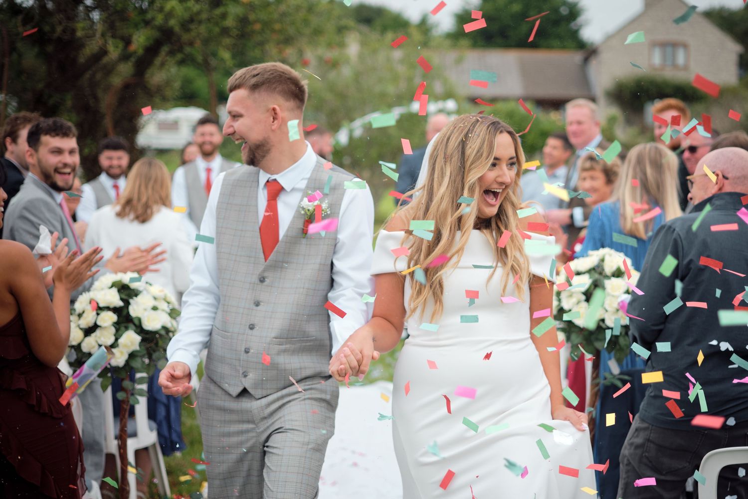 A bride and groom walk through falling confetti petals at their outdoor wedding ceremony as guests celebrate.