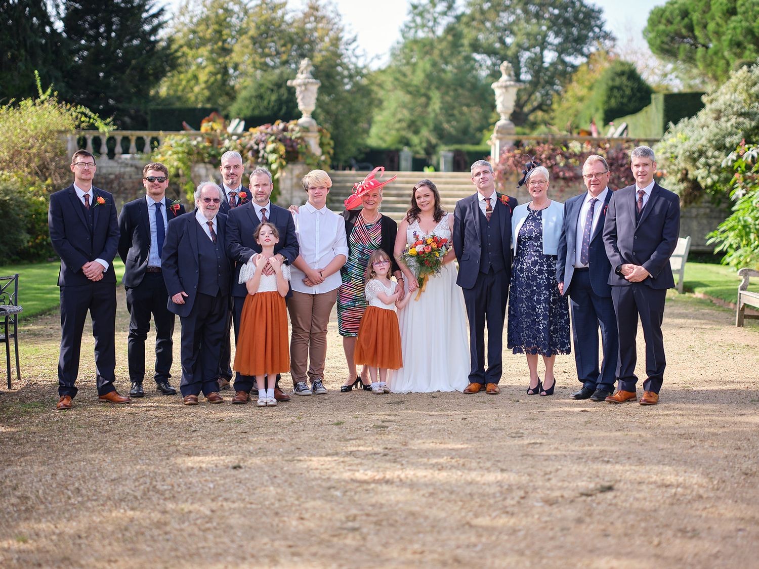 A wedding party poses together outdoors in a garden setting on a sunny autumn day.