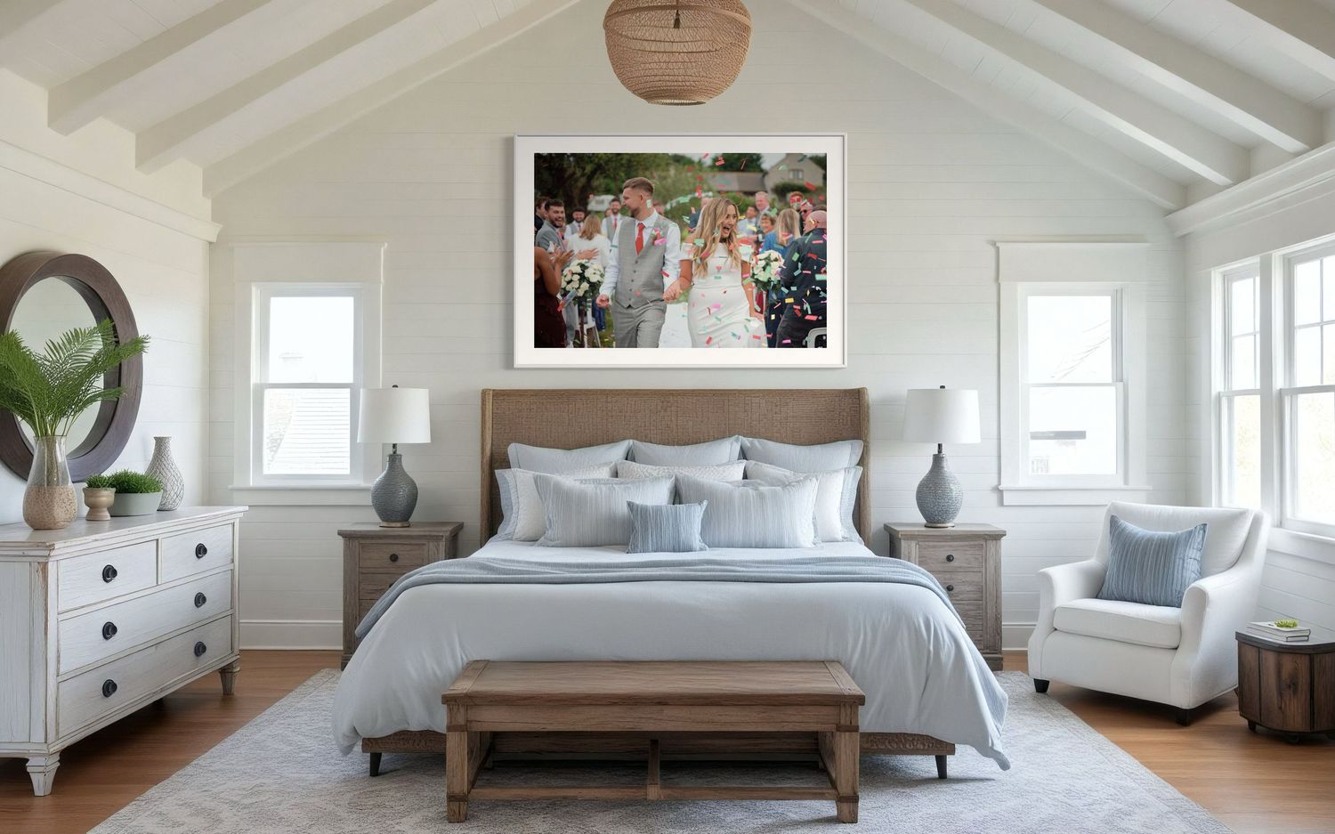 A coastal style bedroom features light blue bedding, white furniture, and vaulted ceiling with a large framed photograph above the bed.