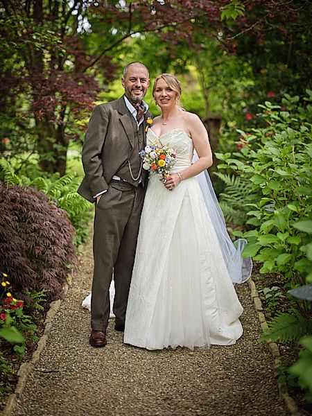 A newly married couple poses together on a garden path surrounded by lush greenery and flowering plants.