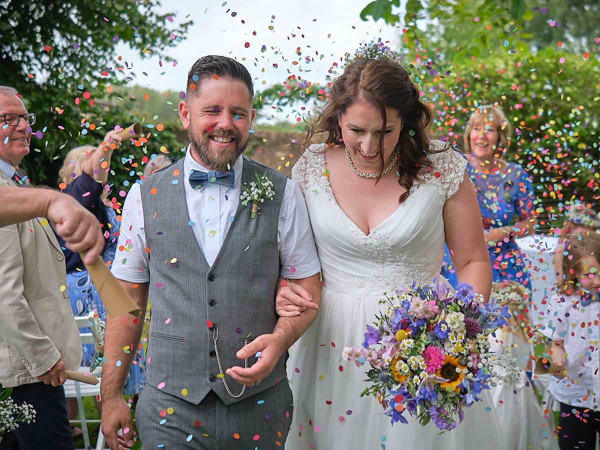 Wedding celebration with confetti in the air as newlyweds walk among guests with colorful wildflower bouquet.