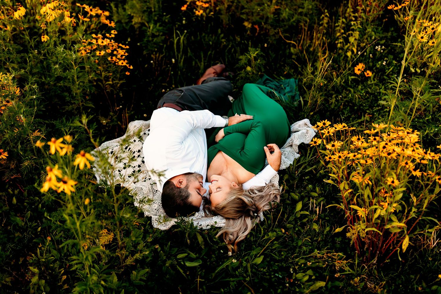 A couple lies on a blanket amidst vibrant yellow wildflowers. They're dressed elegantly, embracing and gazing at each other, evoking romance