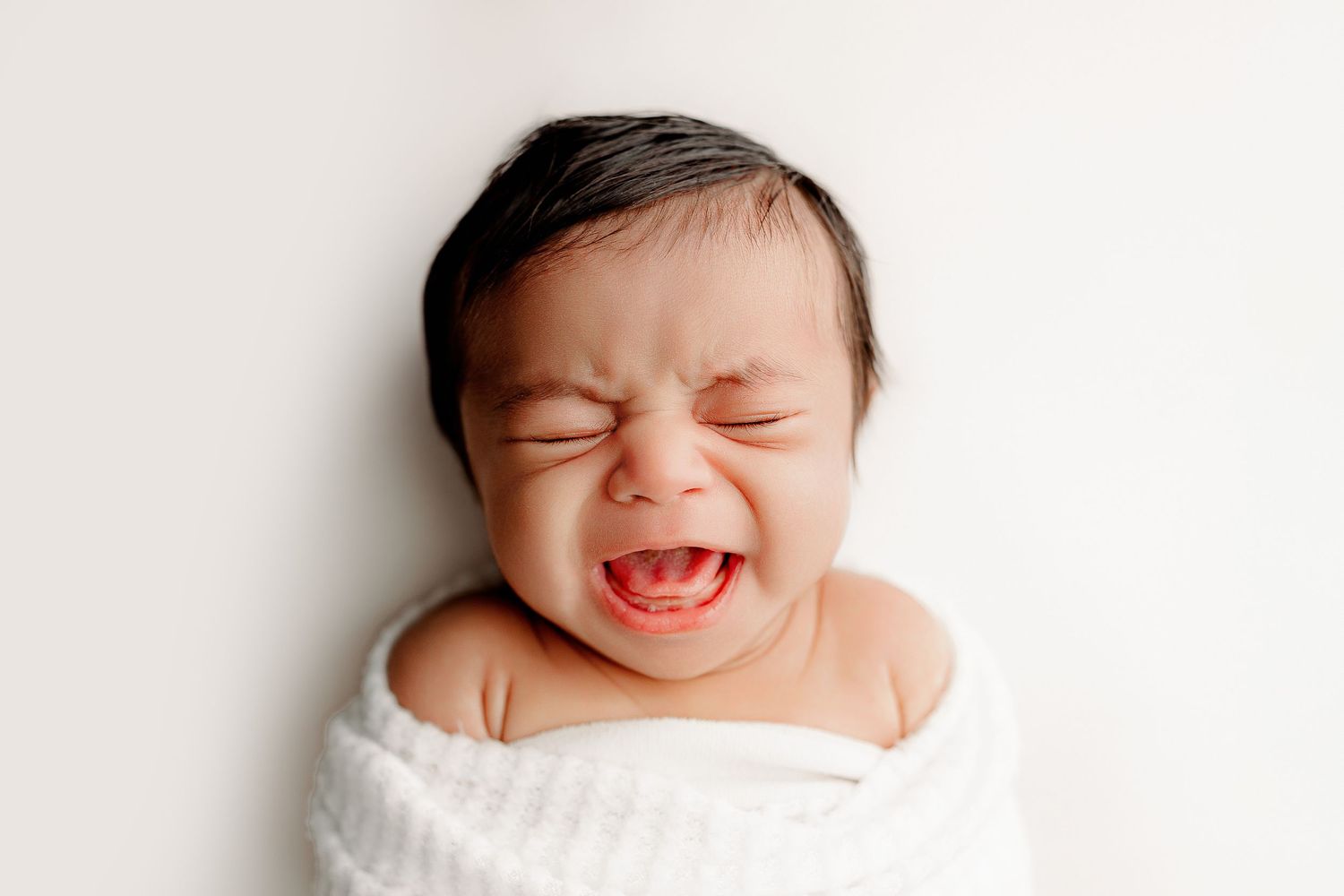 A crying baby wrapped in a soft white blanket lies against a plain background.