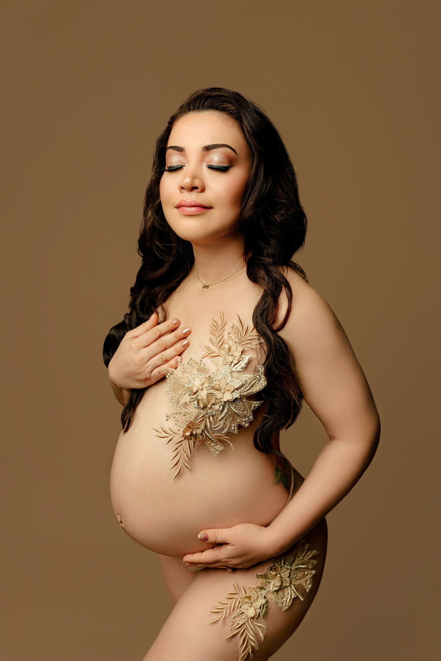 A serene pregnant woman poses against a brown backdrop, adorned with intricate beige floral designs, one hand on her belly