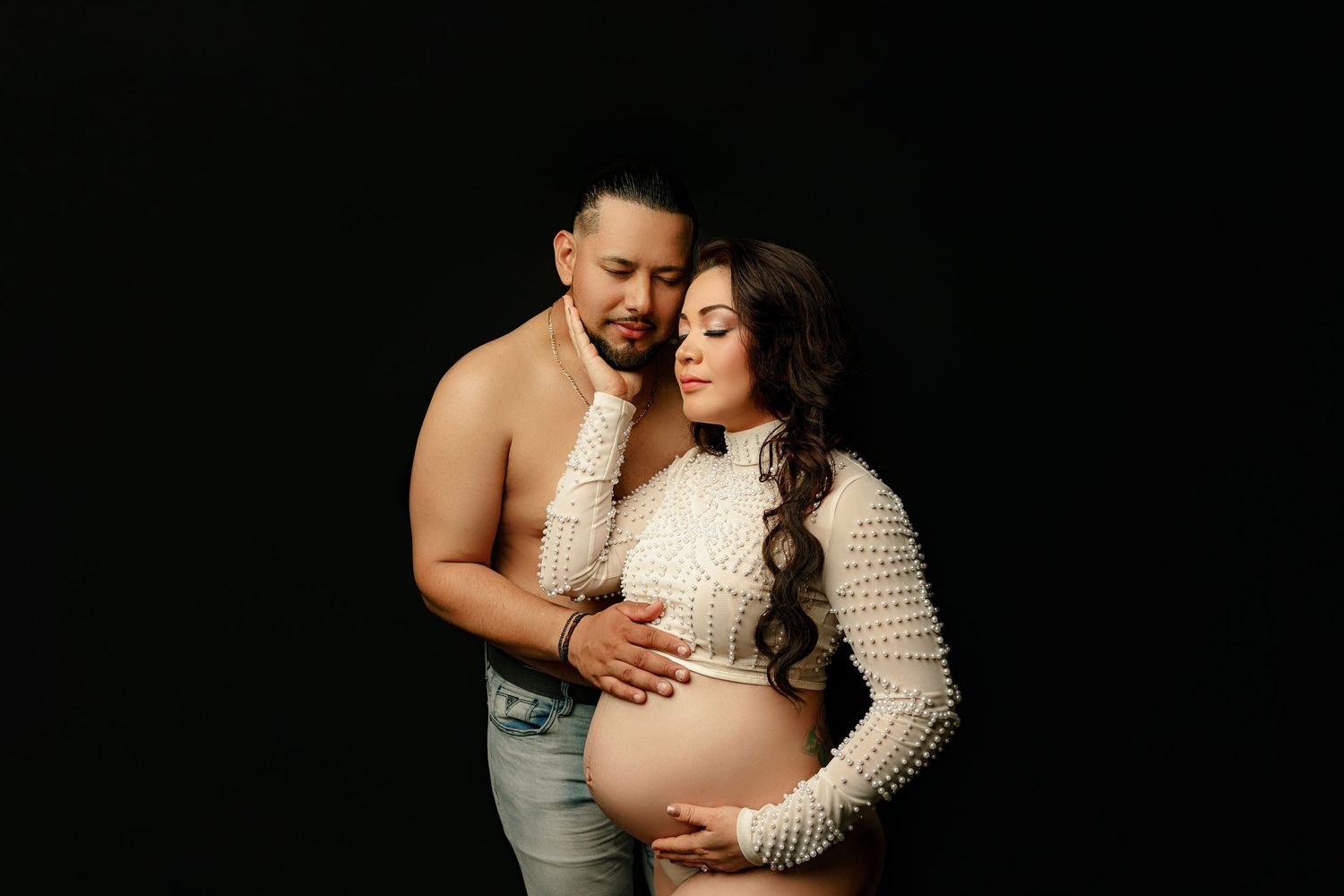 A shirtless man and a pregnant woman in a white textured dress share an intimate moment against a black background