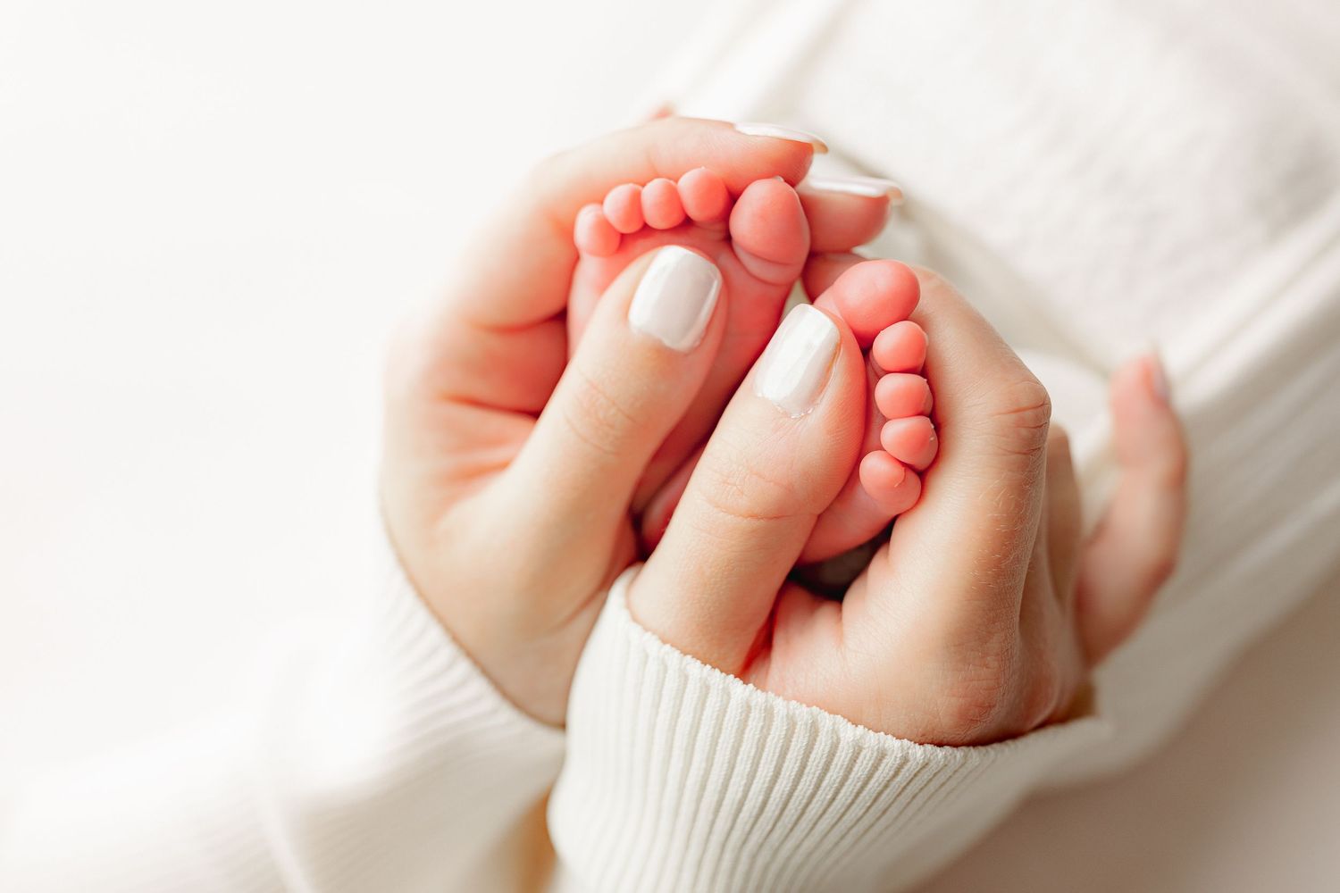 Two adult hands gently cradle tiny baby feet, conveying warmth and tenderness. The adult wears a white sweater