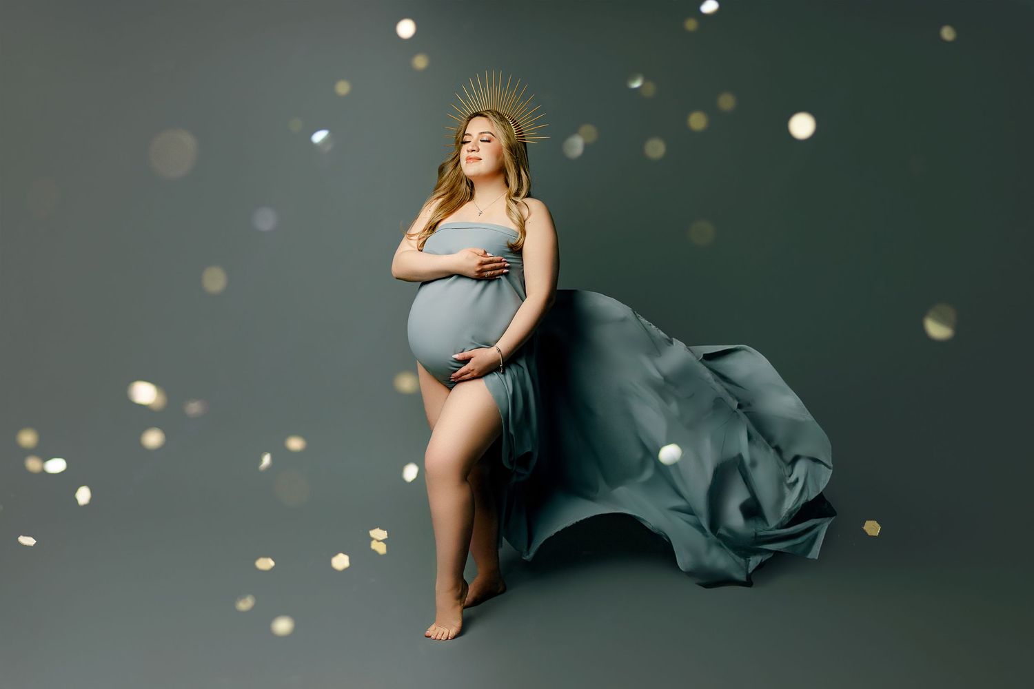 A pregnant woman stands barefoot in a flowing green gown, adorned with a radiant gold halo crown. Soft light creates an ethereal scene