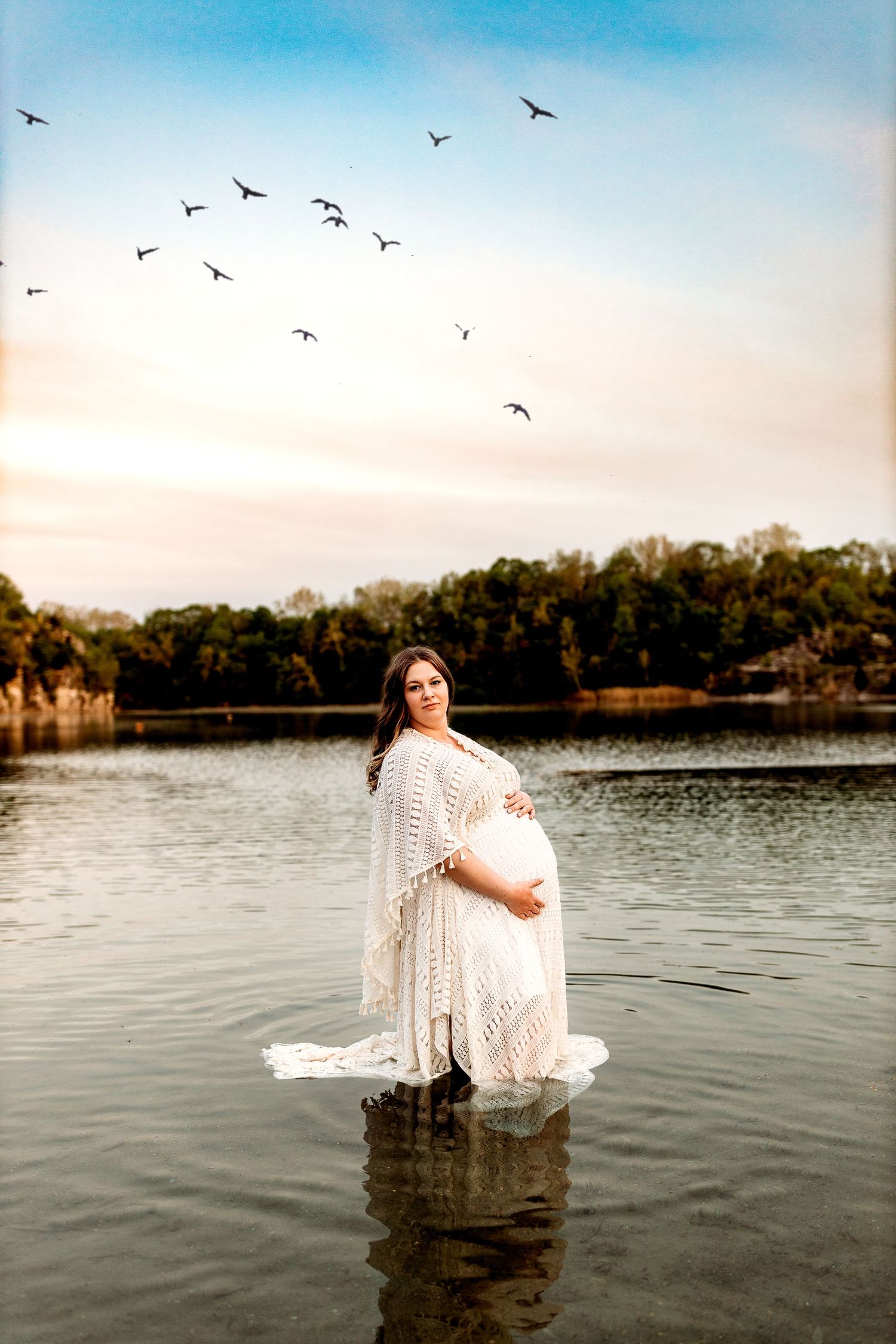 Pregnant woman in a white dress stands in a calm lake, surrounded by a serene landscape with distant trees and a flock of birds in the sky