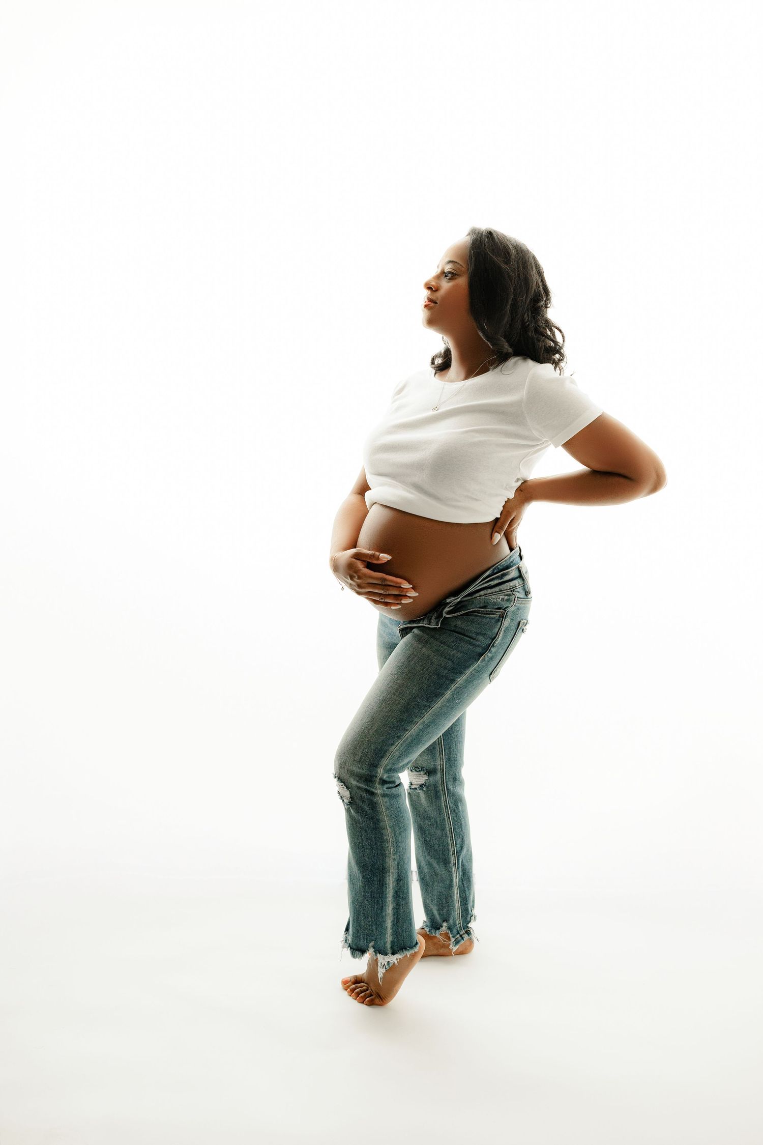 Pregnant woman standing in profile against a white background, wearing a white shirt and jeans, cradling her belly