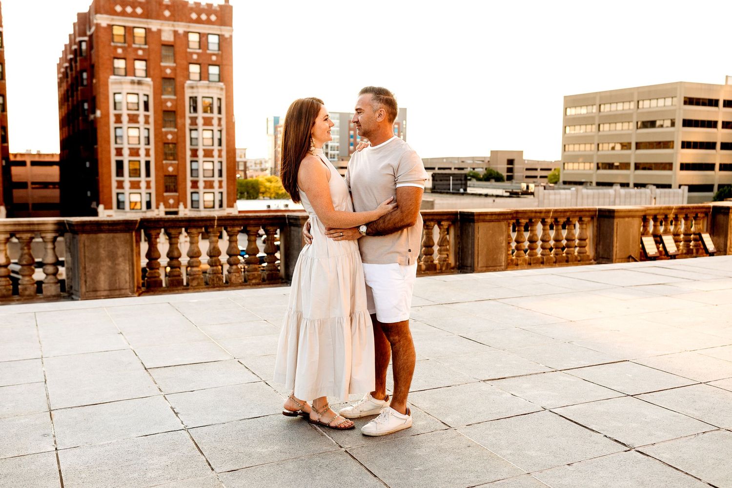 A couple embraces on a city rooftop at sunset. They stand facing each other, surrounded by tall buildings in downtown Indianapolis Indiana