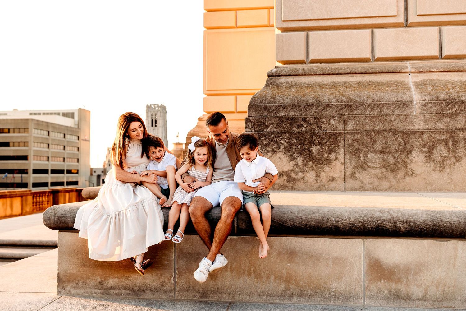 A family of five sits on a stone ledge outdoors, laughing together. The parents and the children are dressed in casually