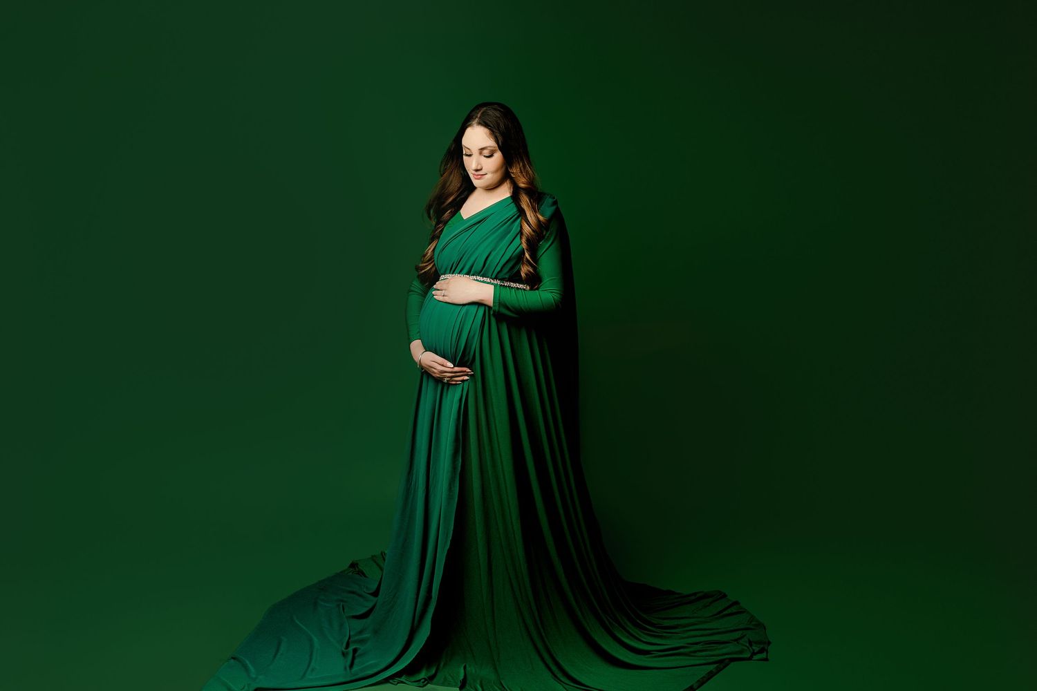 Pregnant woman in a flowing green gown stands against a matching backdrop, gently cradling her belly.