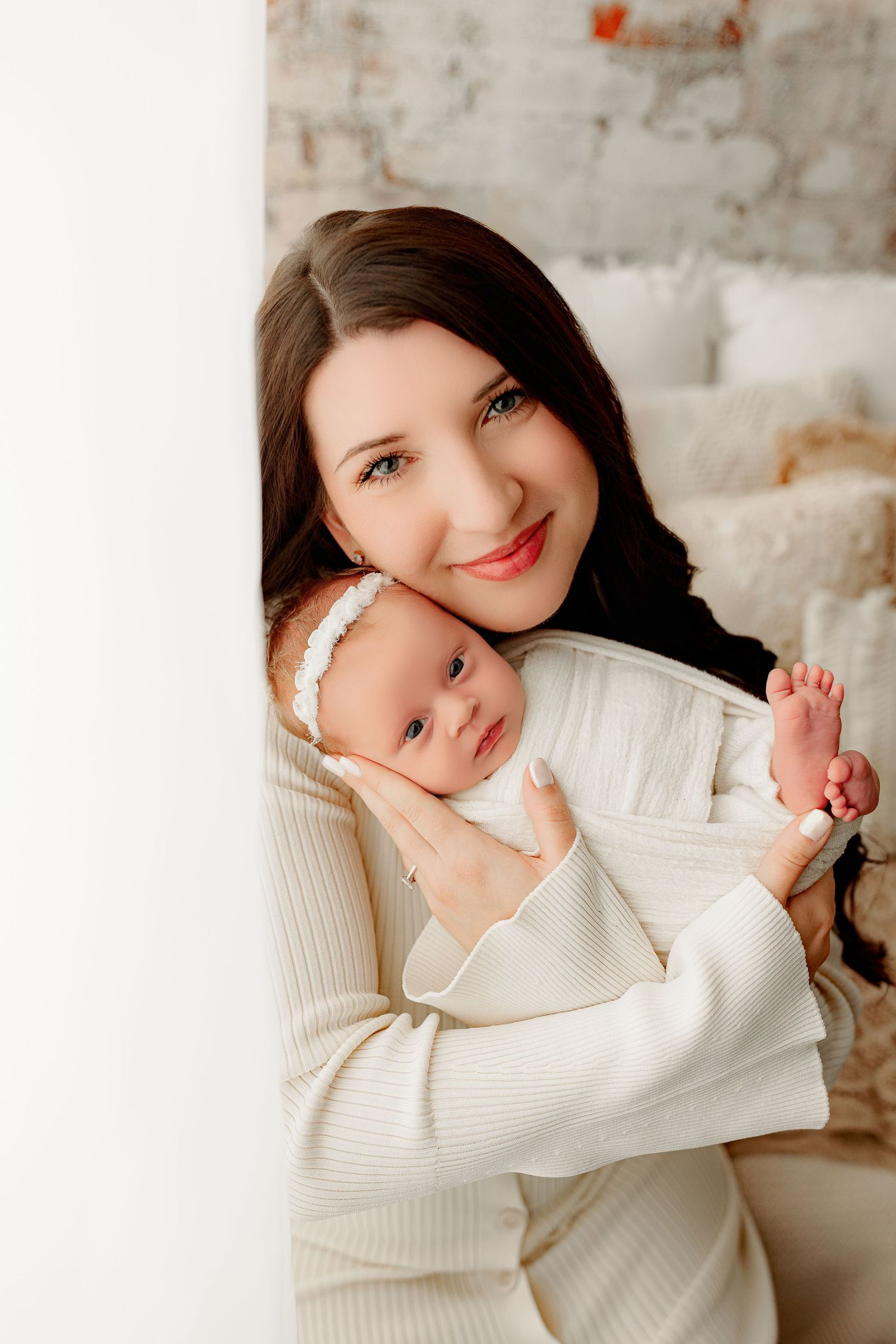 A woman in a white sweater smiles warmly, holding a swaddled baby with a white headband. They sit in a soft, cozy room with neutral tones.