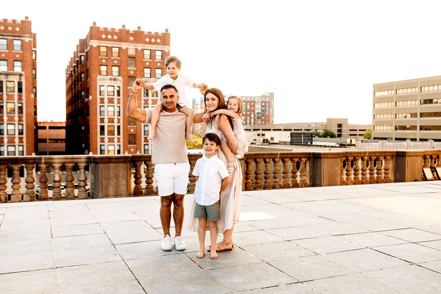 A family of four stands on a rooftop at sunset, with parents carrying two children on their shoulders. They are smiling, with city buildings