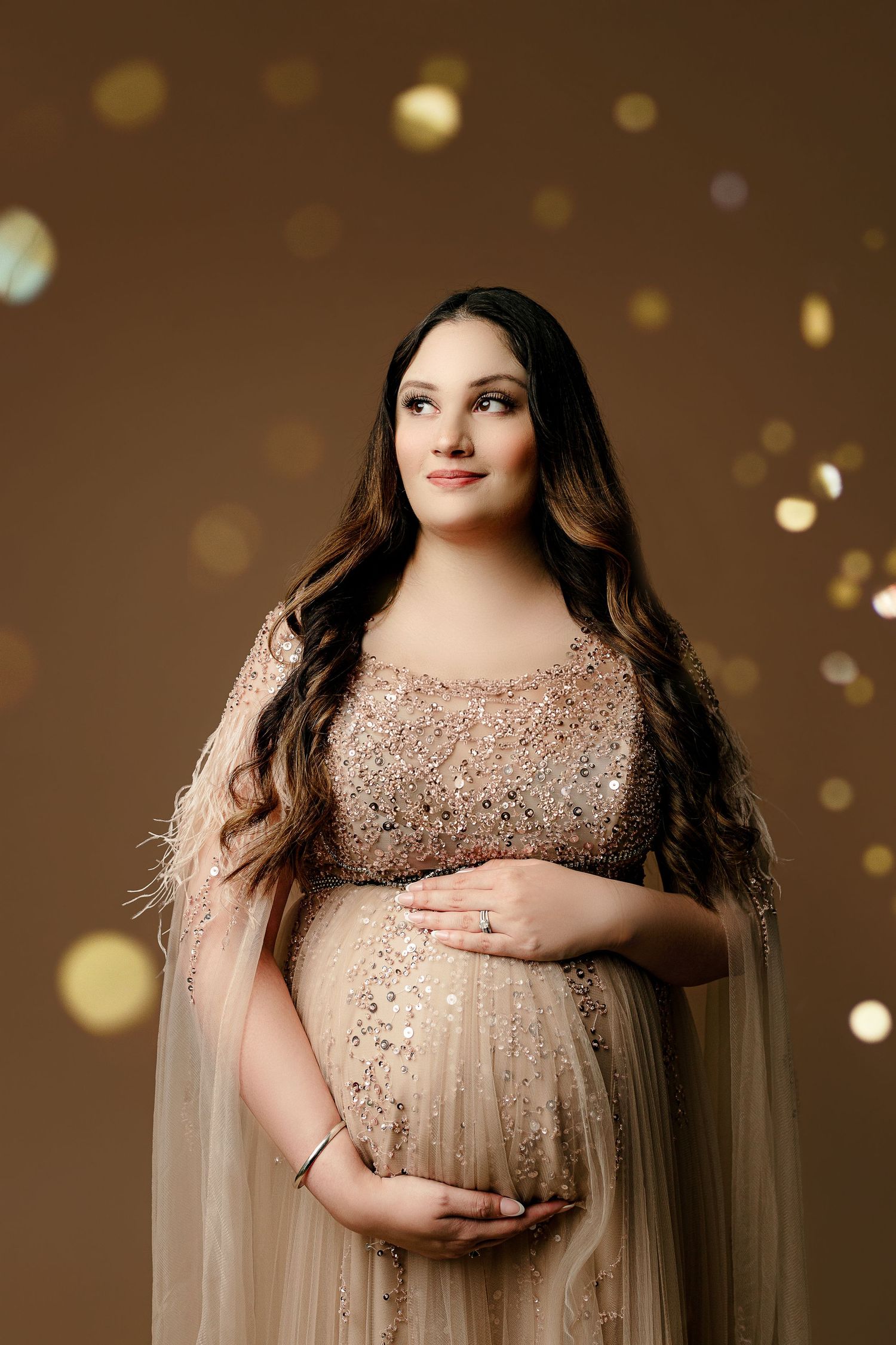 Pregnant woman in a sparkling, beige gown cradles her belly against a brown, bokeh-lit background, exuding warmth and anticipation.