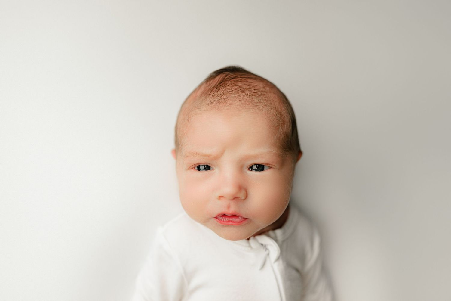 A newborn baby wearing a white outfit rests against a soft, white background. The baby has a curious expression, with furrowed brows