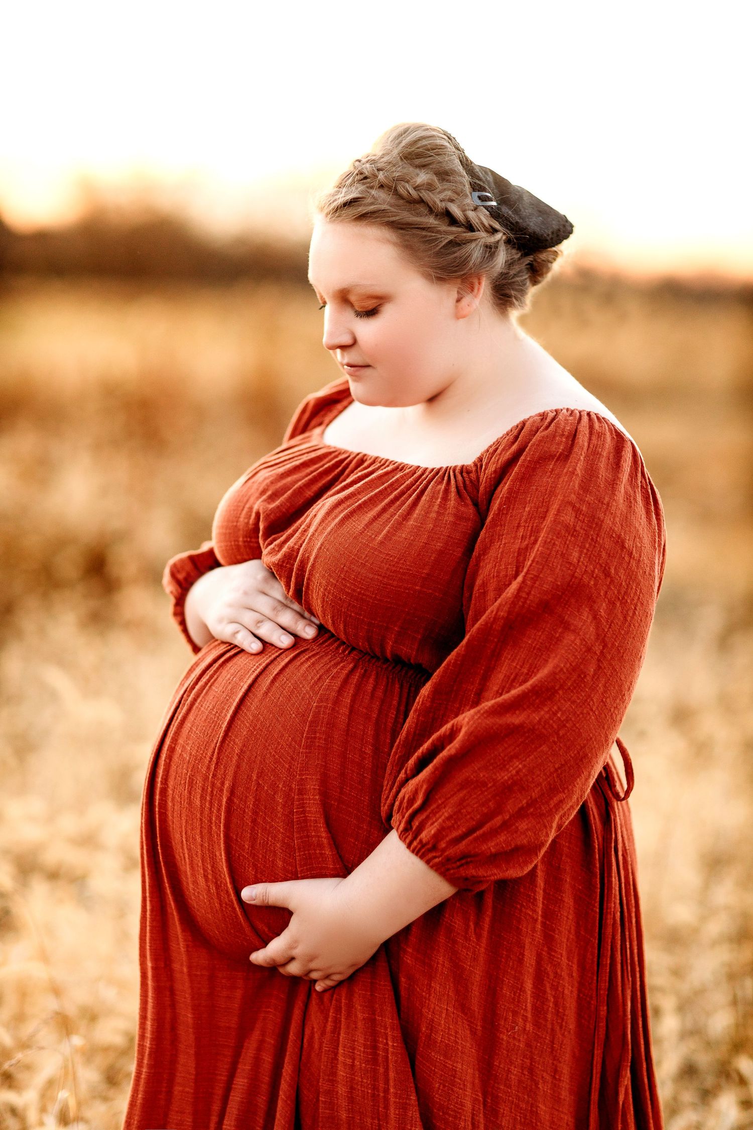 A pregnant woman in a flowing, rust-colored dress stands in a sunlit wheat field, gently cradling her belly. The scene conveys warmth
