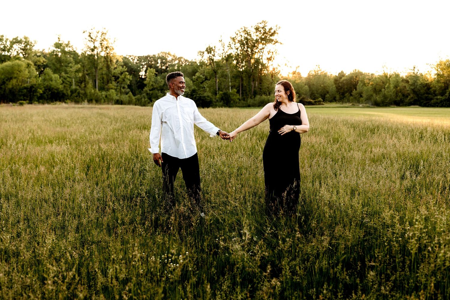 A couple holds hands, walking through a sunlit meadow. The man wears a white shirt; the woman, a black dress. Both smile in a green field