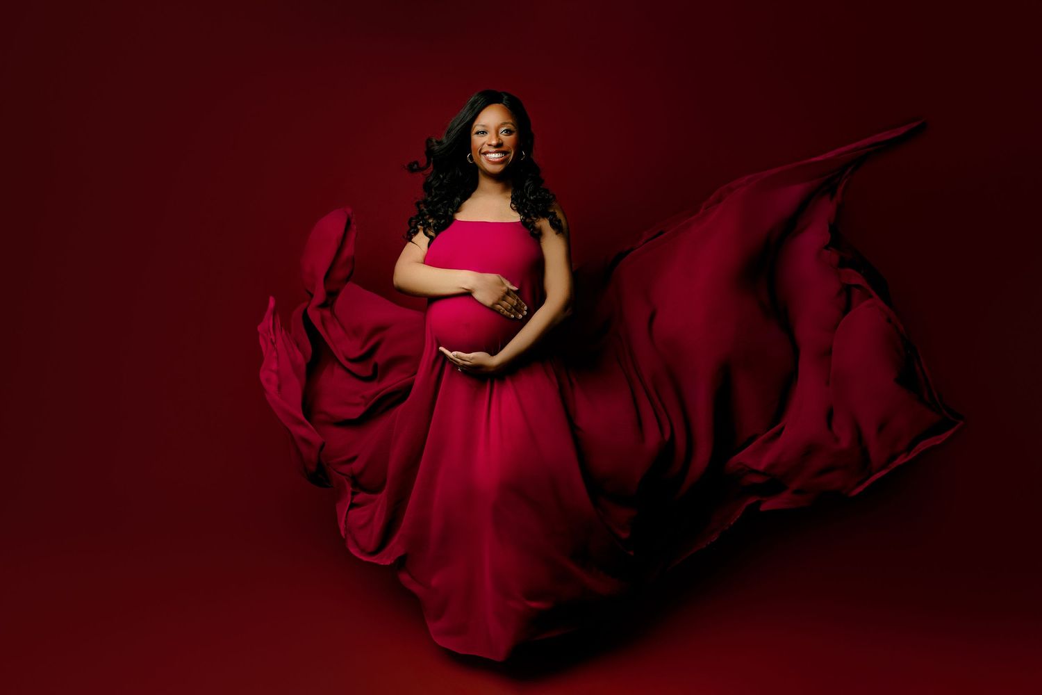 A smiling pregnant woman in a flowing red gown poses against a dark red background. The dress billows around