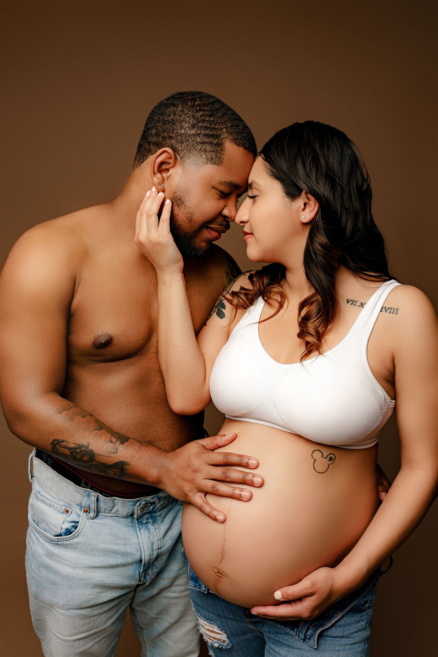 A couple embraces tenderly against a brown background. The man is shirtless, and she cradles her pregnant belly wearing a white top and jean