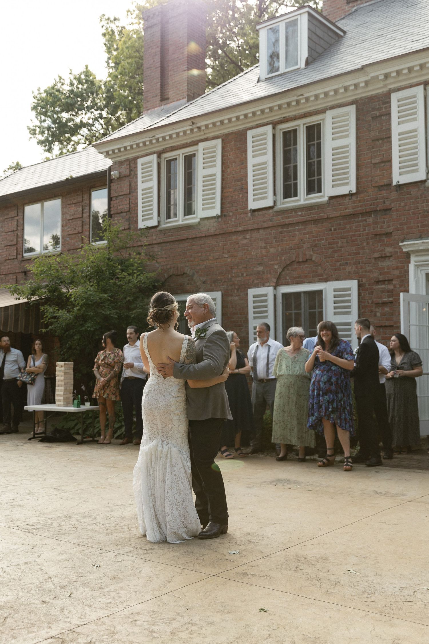 A couple sharing their first dance at an outdoor wedding venue with brick building and guests watching in background.