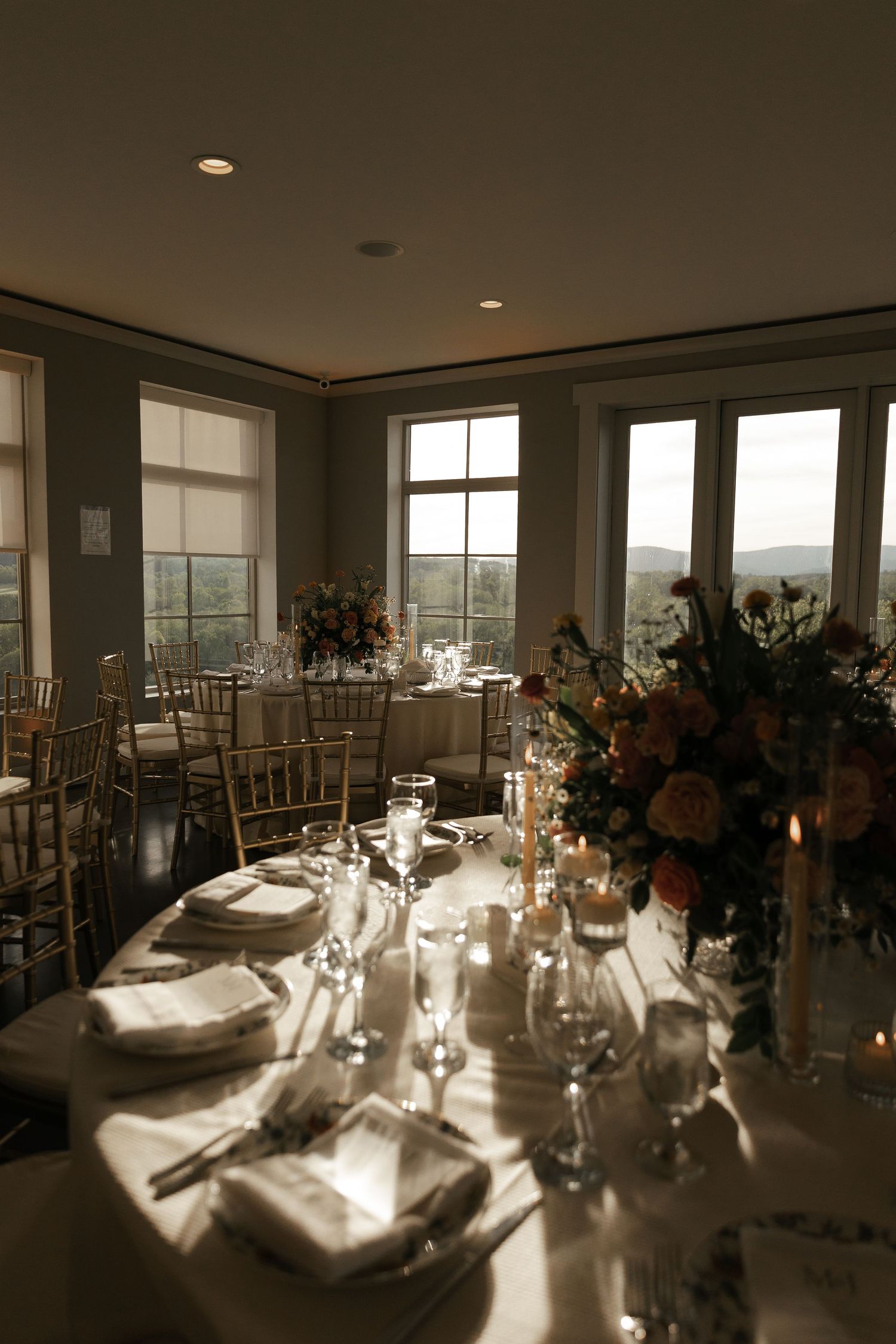 Elegant wedding reception table setting with floral centerpieces and sunlight streaming through large windows overlooking mountains.