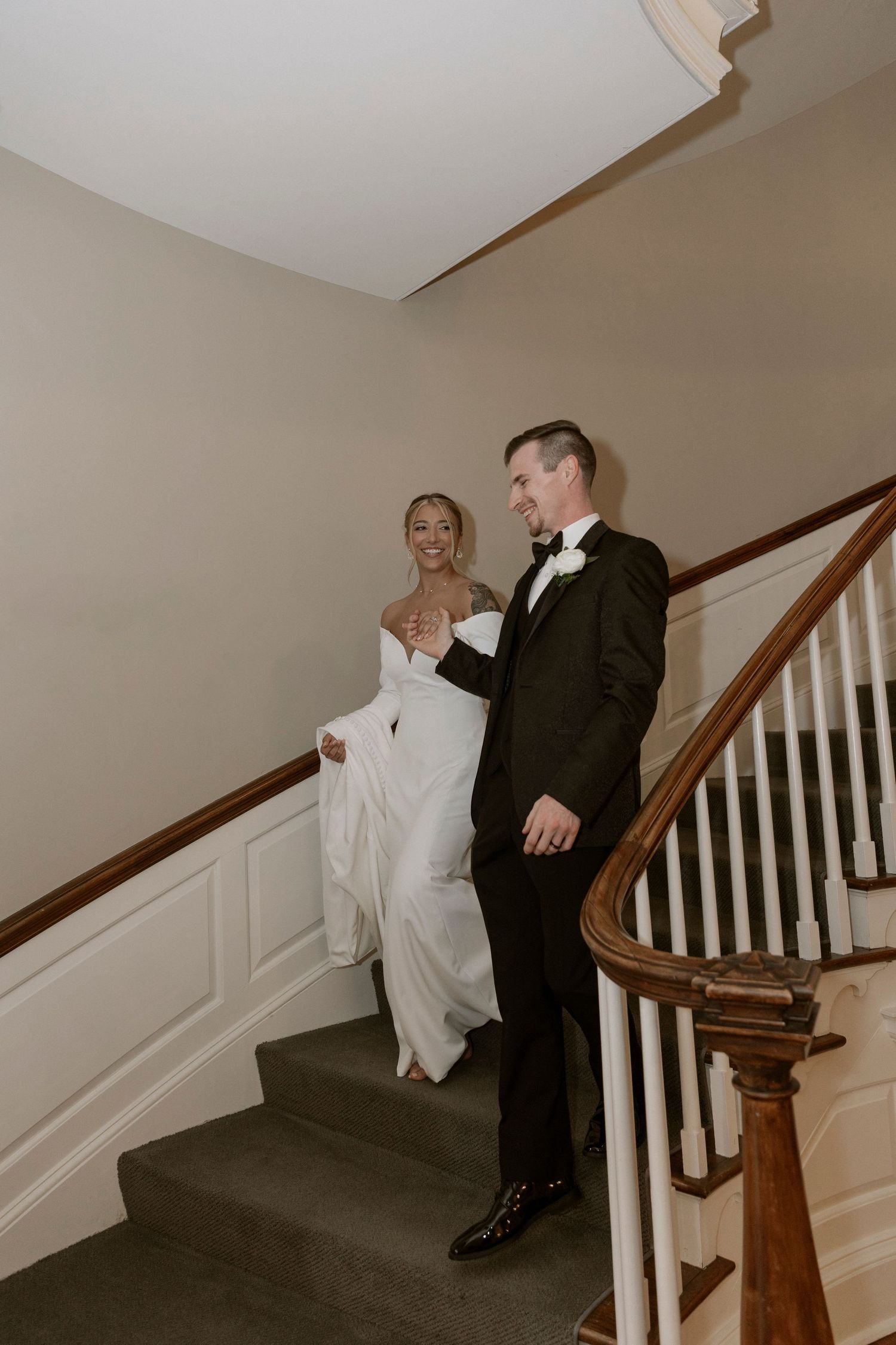 A newlywed couple descends an elegant carpeted staircase with white wainscoting and wooden bannister in warm lighting.