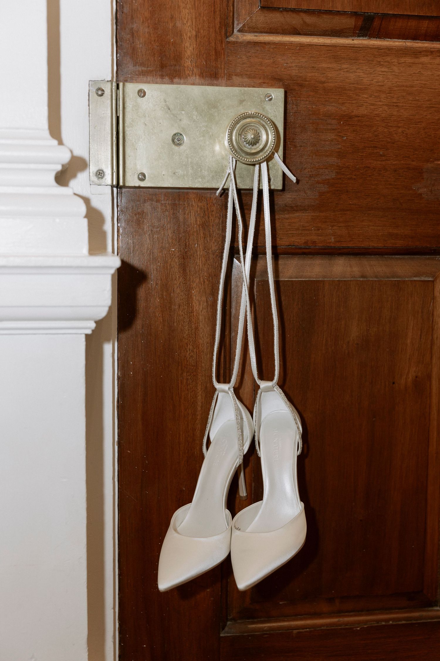 A pair of elegant white high-heeled shoes hanging by their straps from a vintage door lock on a wooden door.
