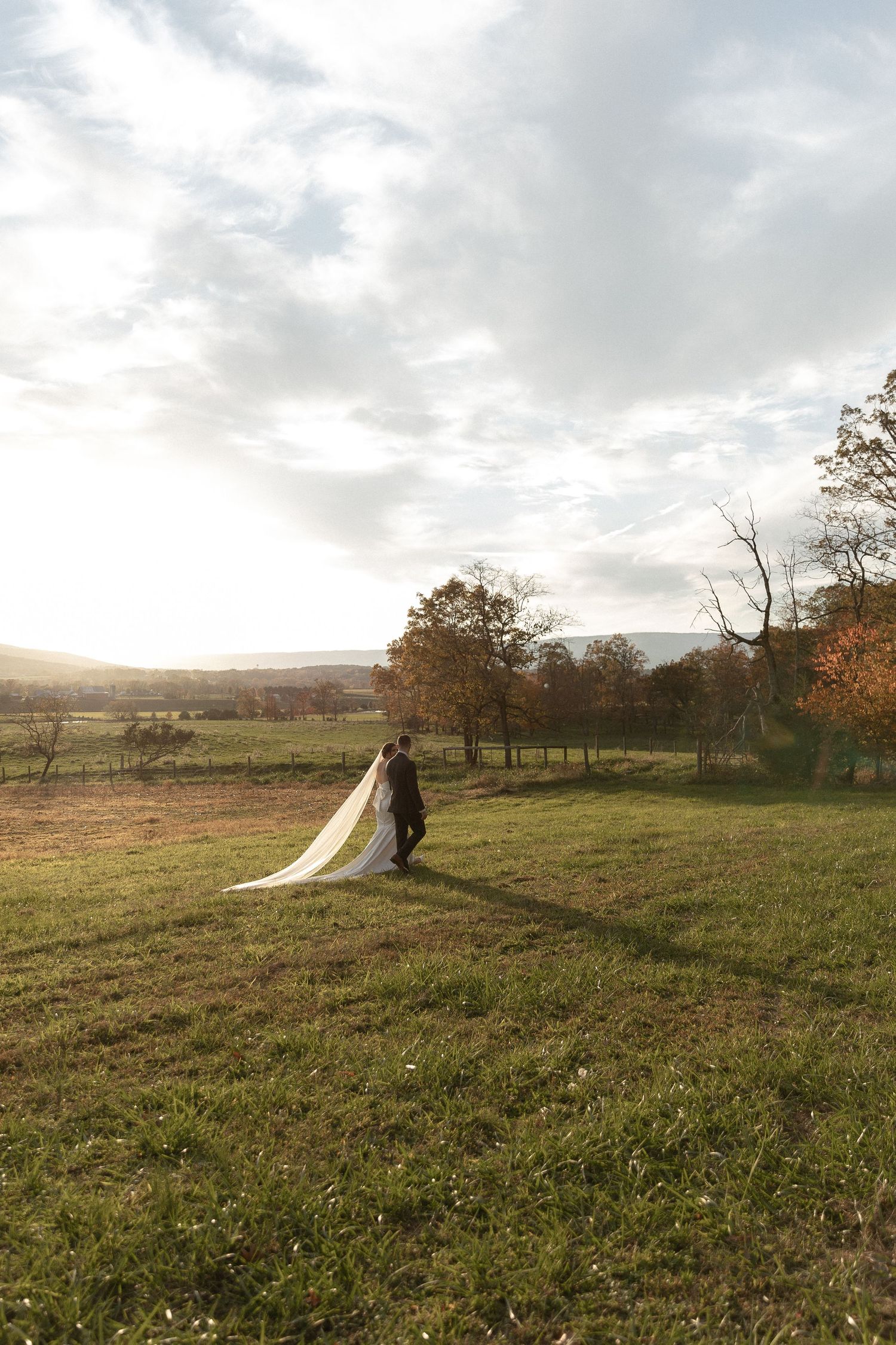 A couple in wedding attire walk across a golden autumn meadow at sunset with rolling hills in the background.