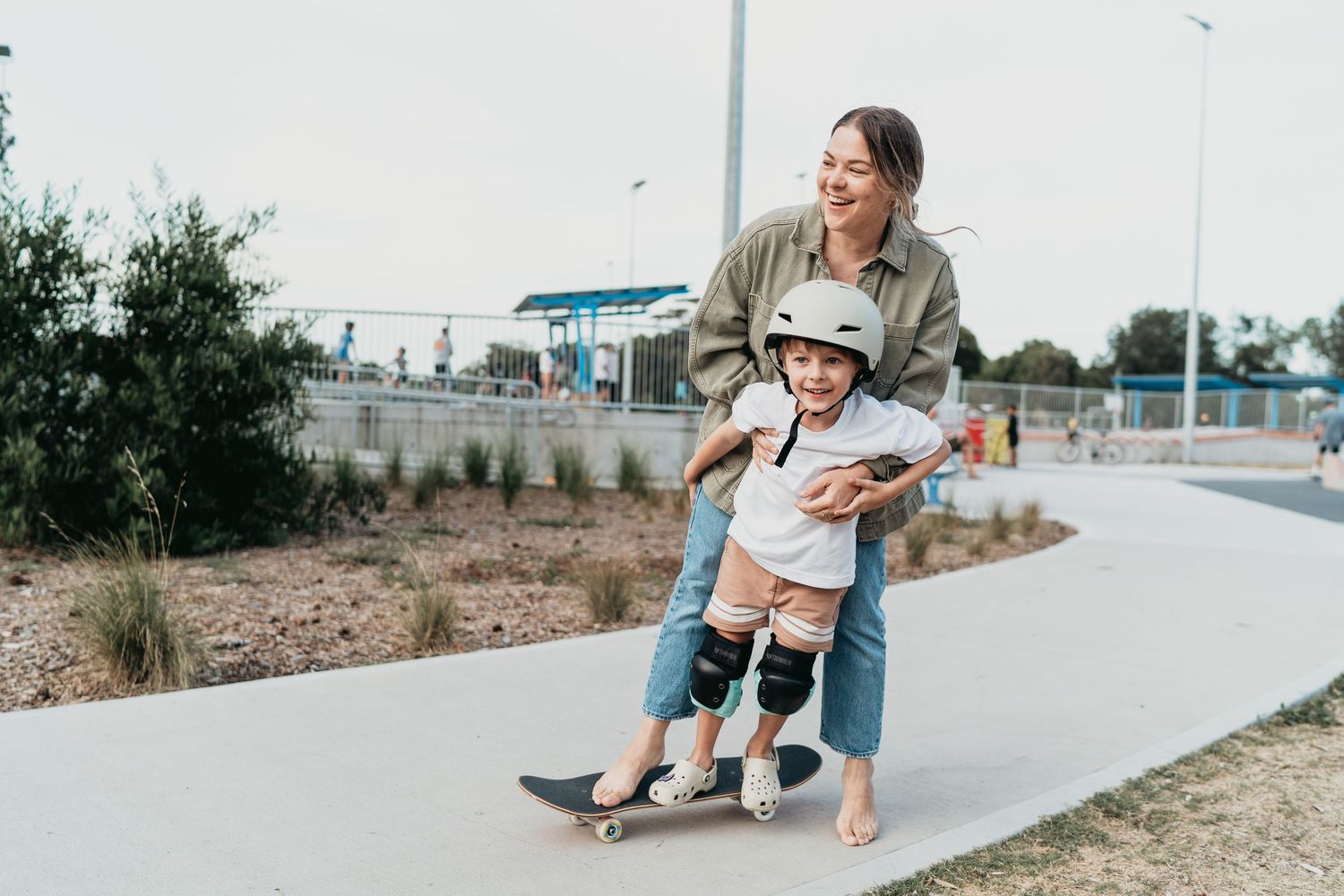 A caregiver helps guide a child learning to skateboard on a paved path near a beach.