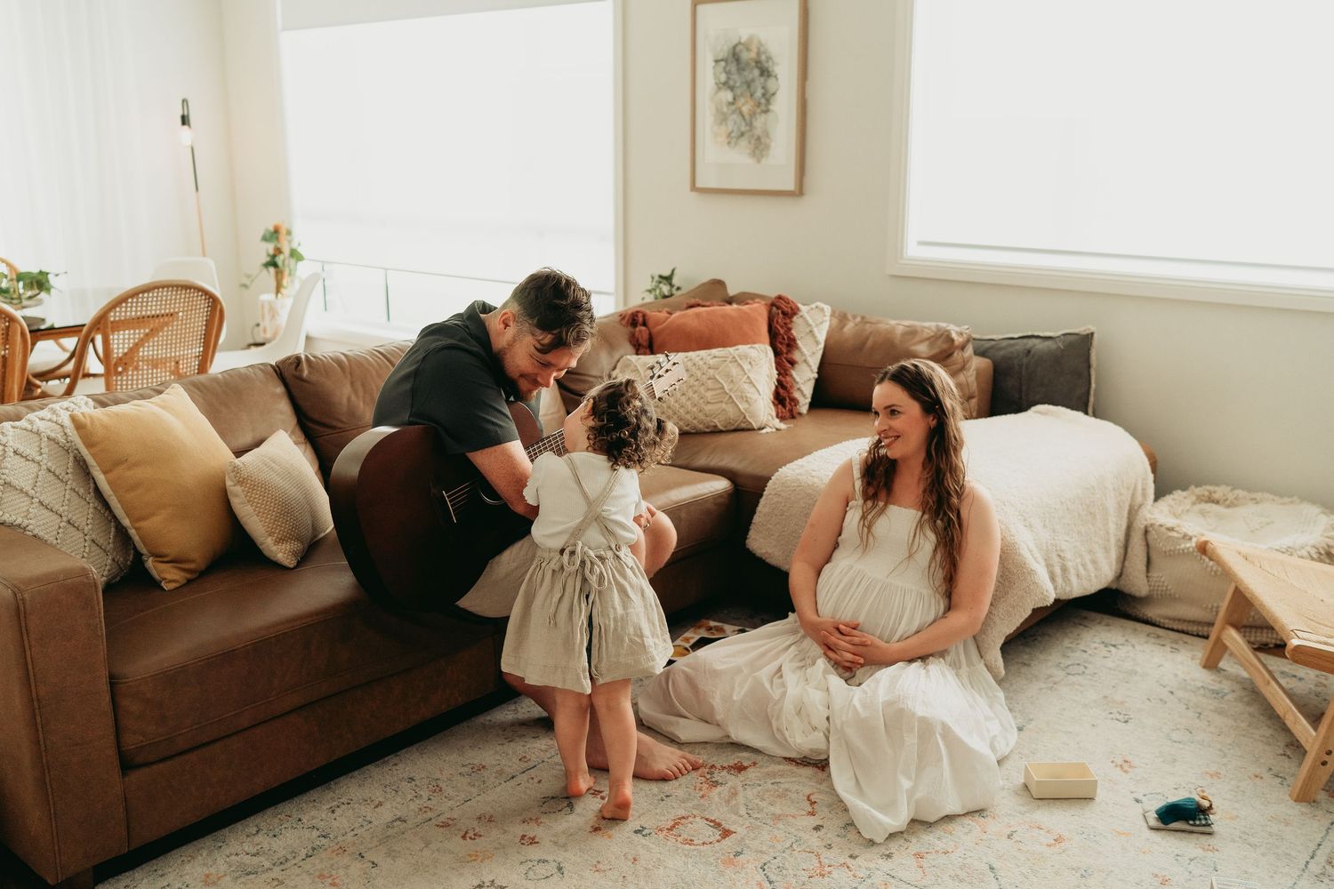 People dressed in white sitting and lying on brown sofas in a bright living room with natural light coming through windows.