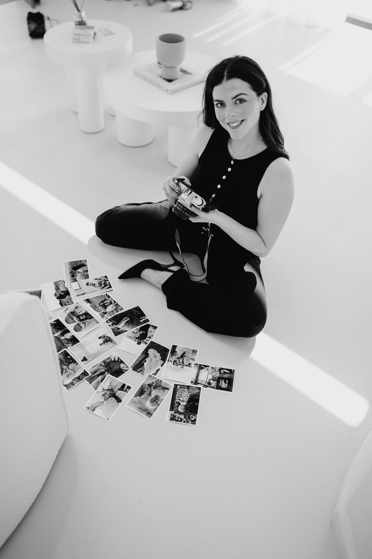 Black and white photo of someone in dark clothing sitting on floor with photographs spread out, holding a camera.