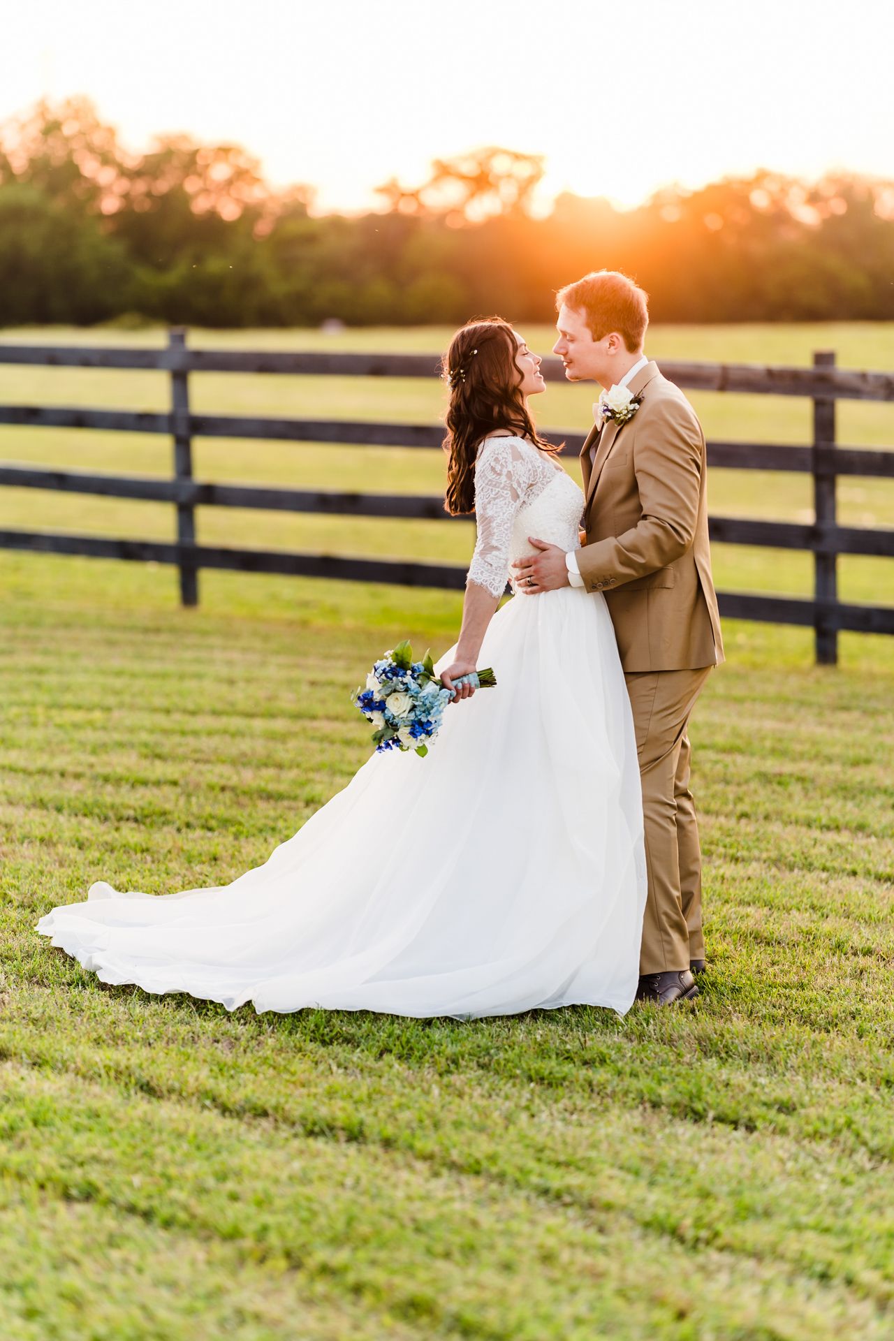 Romantic wedding couple embracing in golden sunset light on farm field with rustic wooden fence in background.