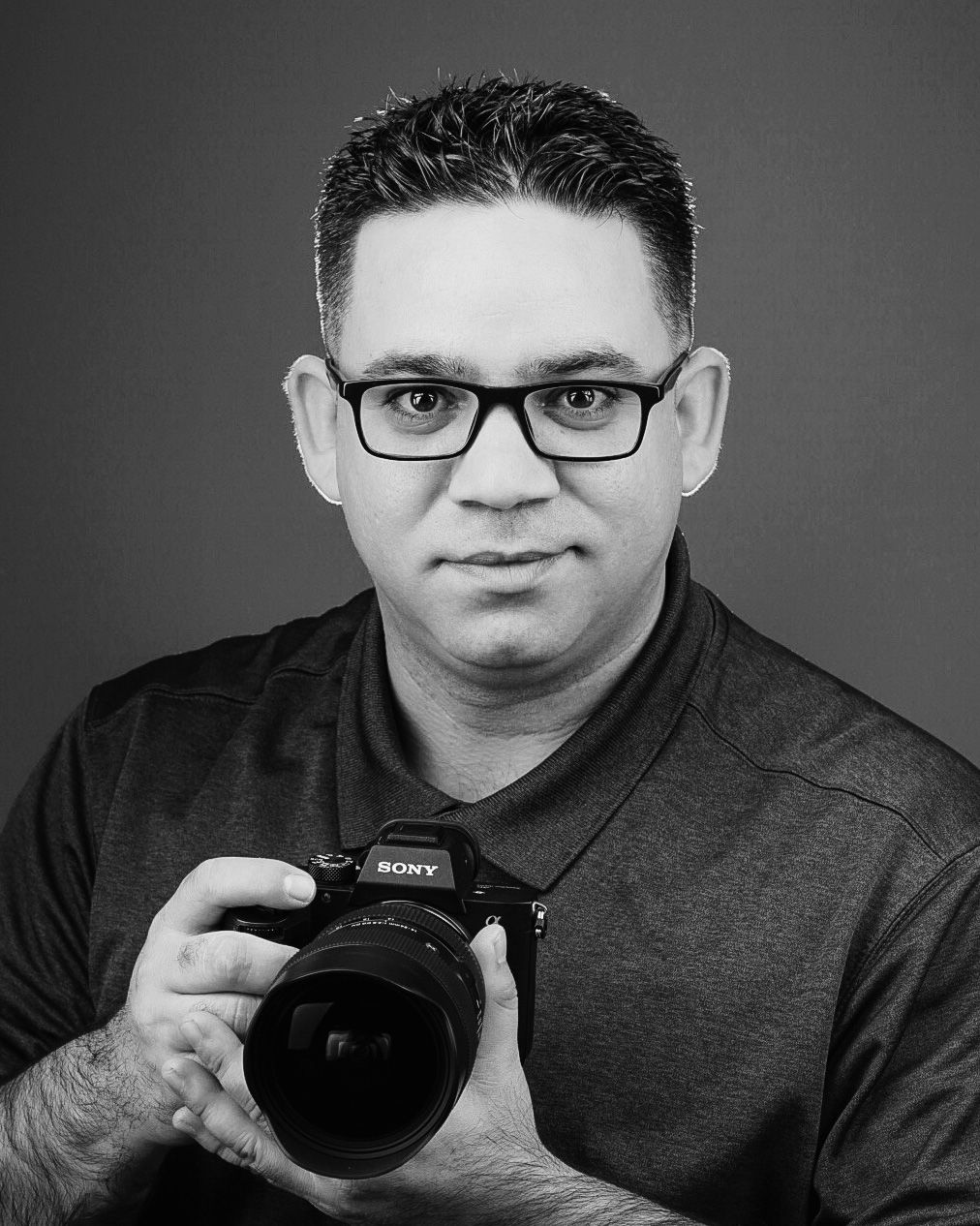 Professional headshot of a photographer wearing glasses and holding a Sony camera against a dark background.