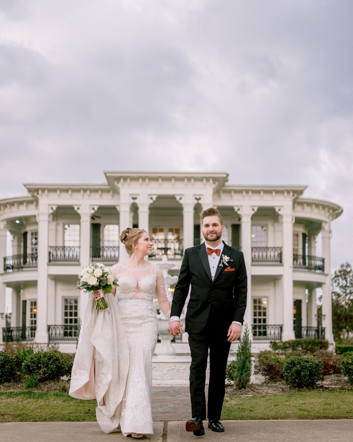 A couple stands in front of a grand antebellum mansion with white columns and ornate architecture on their wedding day.