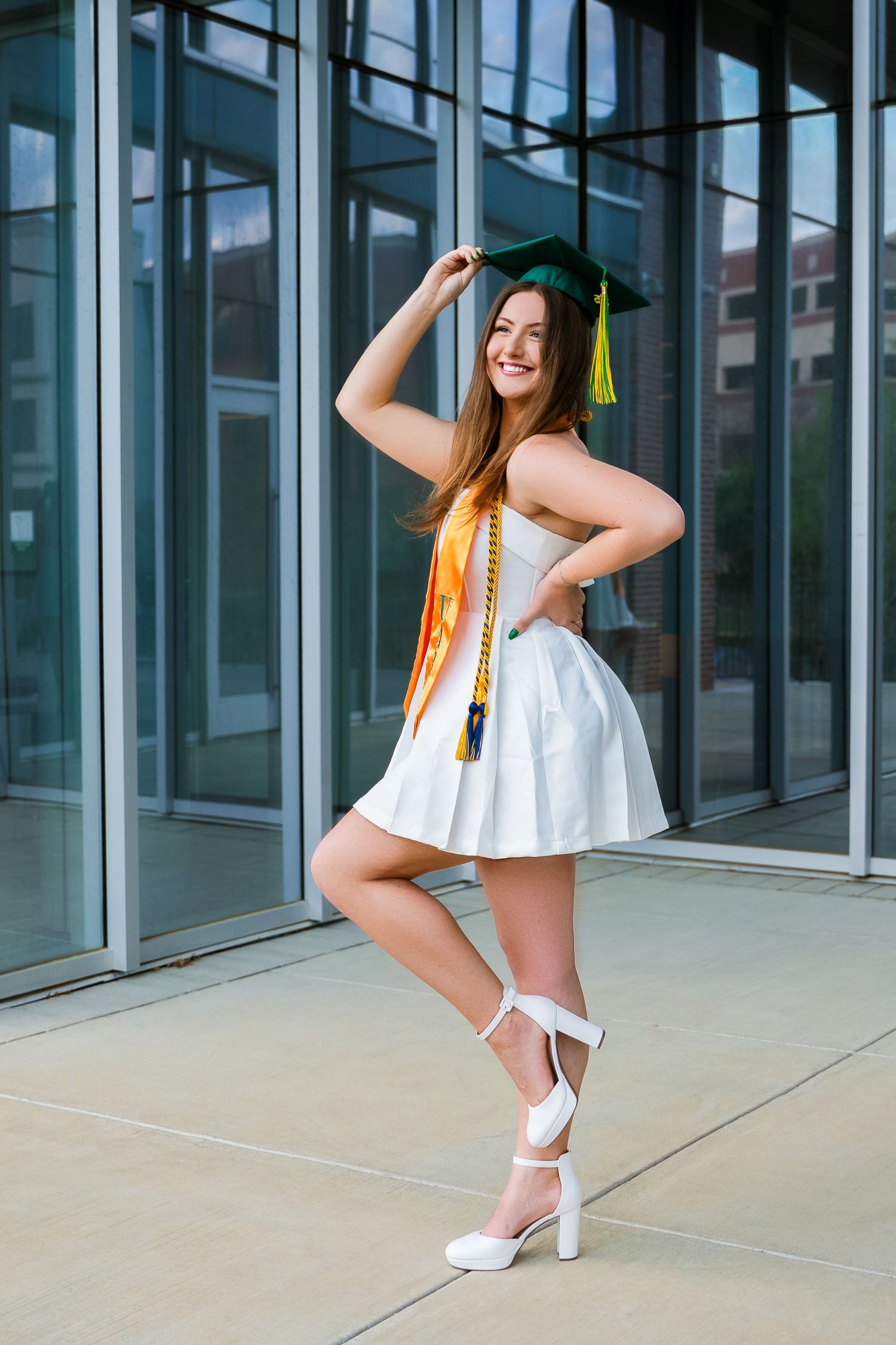 Graduate in white dress poses playfully with graduation cap and tassel.