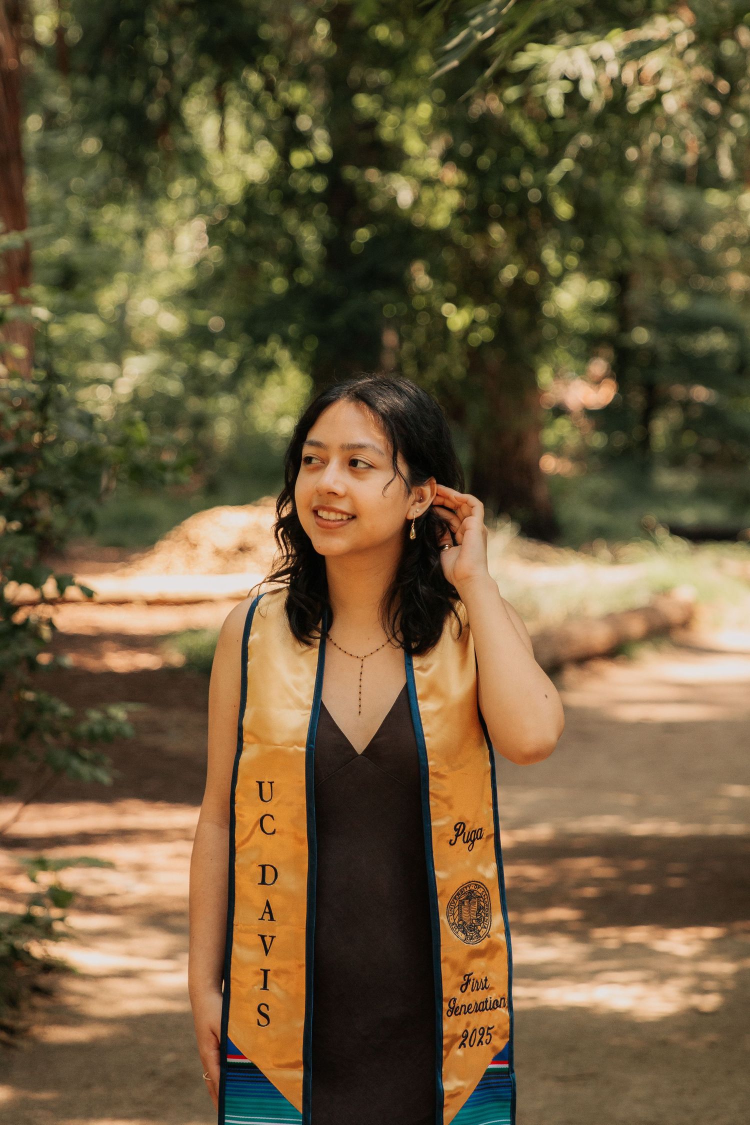 Graduate wearing gold and black UC Davis graduation stole smiles in a sunlit forest setting.