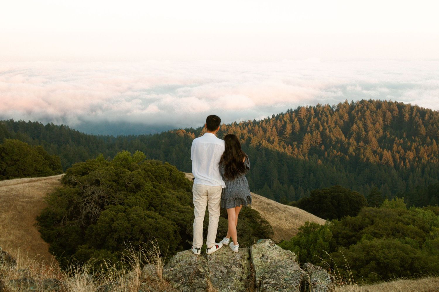 A couple stands at the edge of a mountain overlook taking in sweeping views of forested hills.