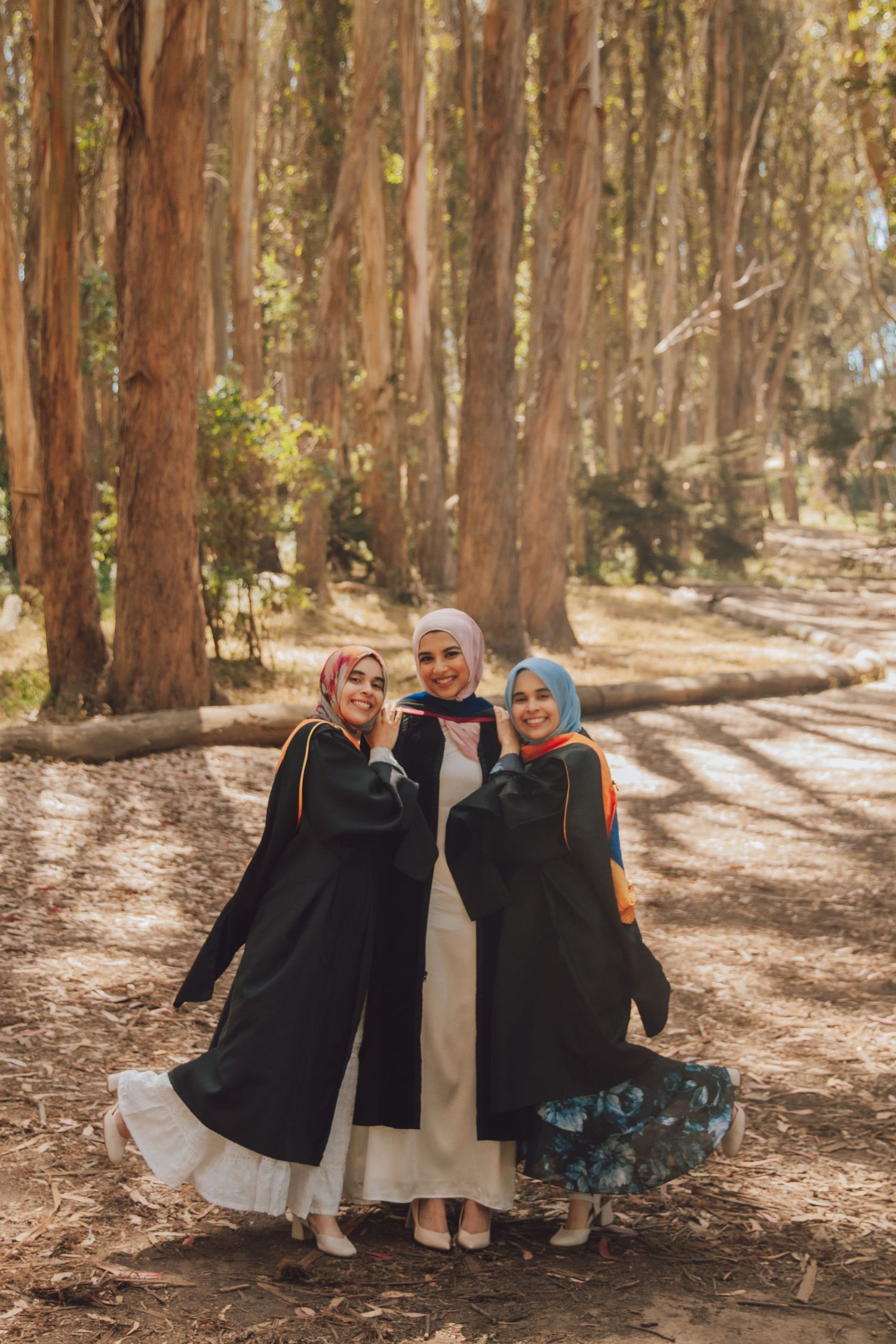Three friends in long modest dresses and headscarves stand together on a sunlit forest path among tall trees.