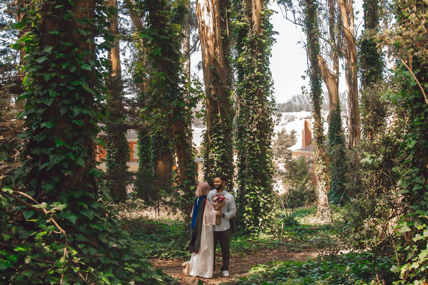 A romantic couple embraces in a mystical forest setting with tall trees wrapped in climbing ivy and dappled sunlight.
