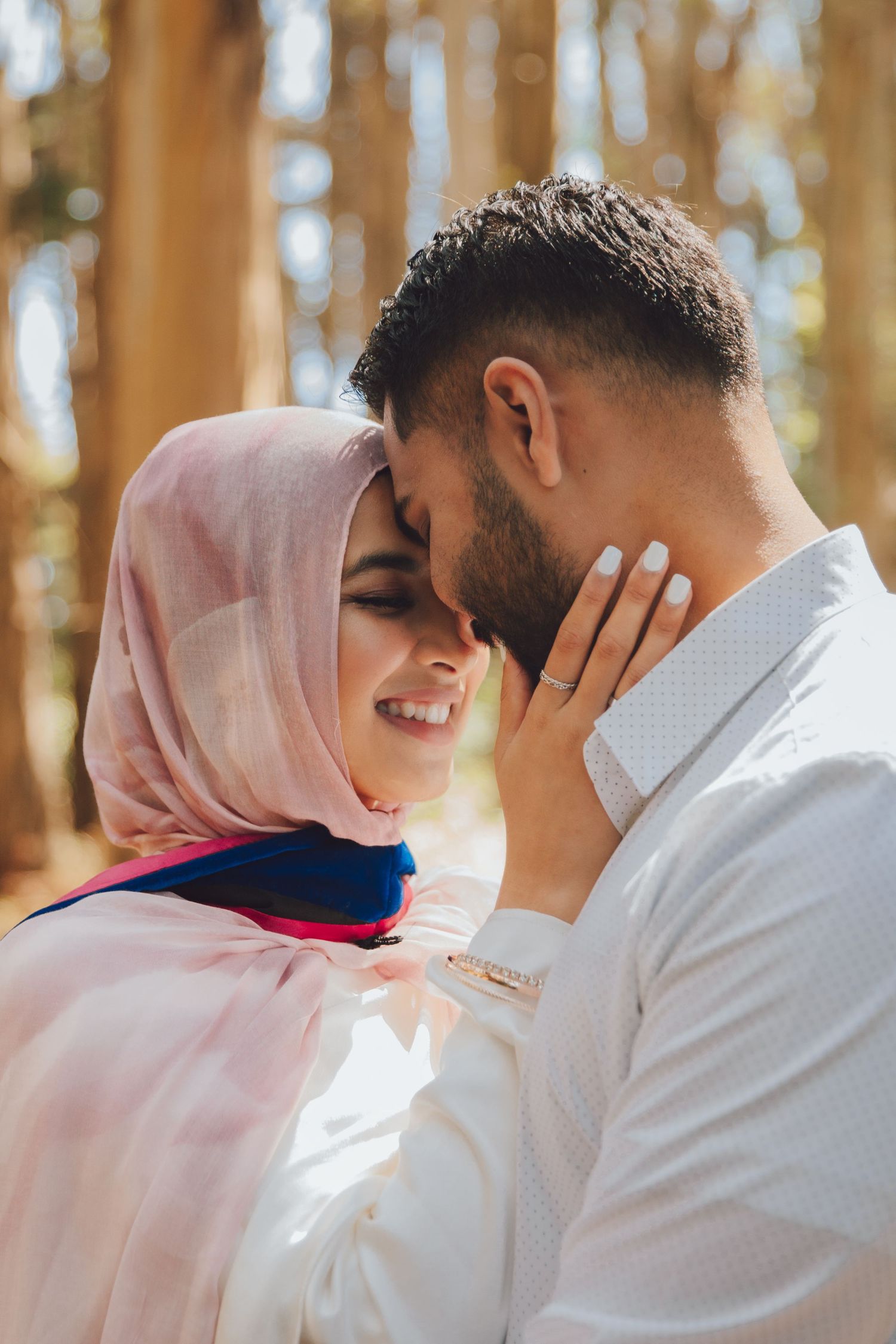 Couple sharing an intimate moment in sunlit forest setting with woman wearing pink hijab and man in white shirt.