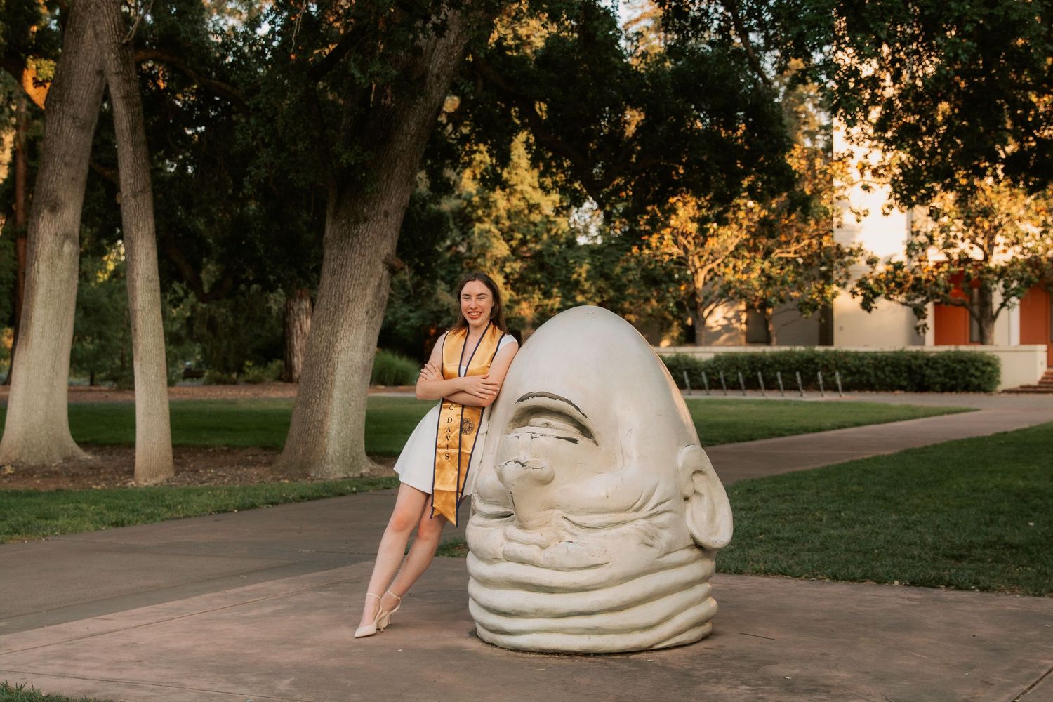 A person sits contemplatively beside a large decorative stone sculpture in a serene outdoor park setting at sunset.